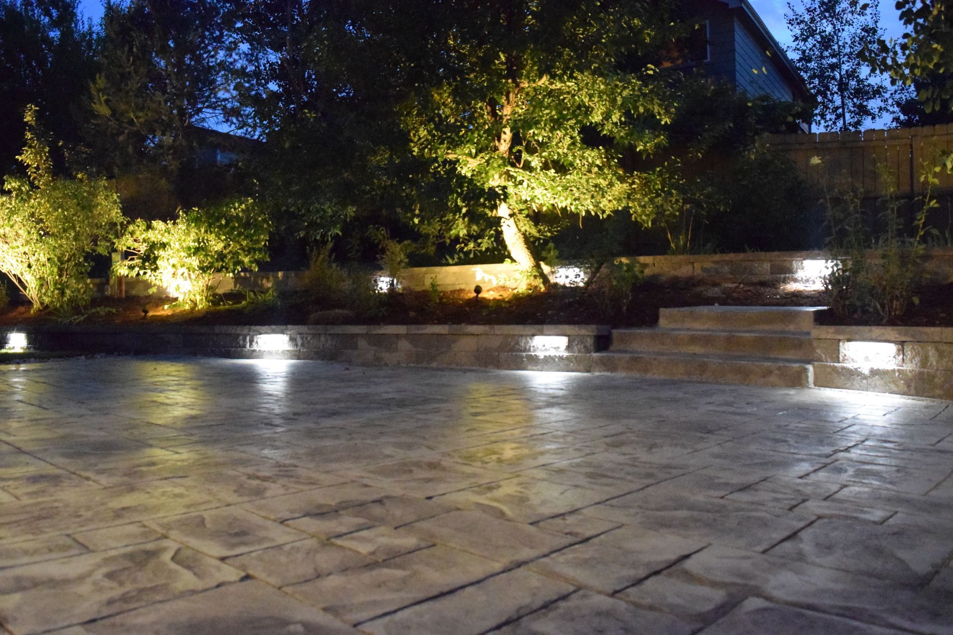 Nighttime outdoor scene with lit patio, retaining walls, and tree.