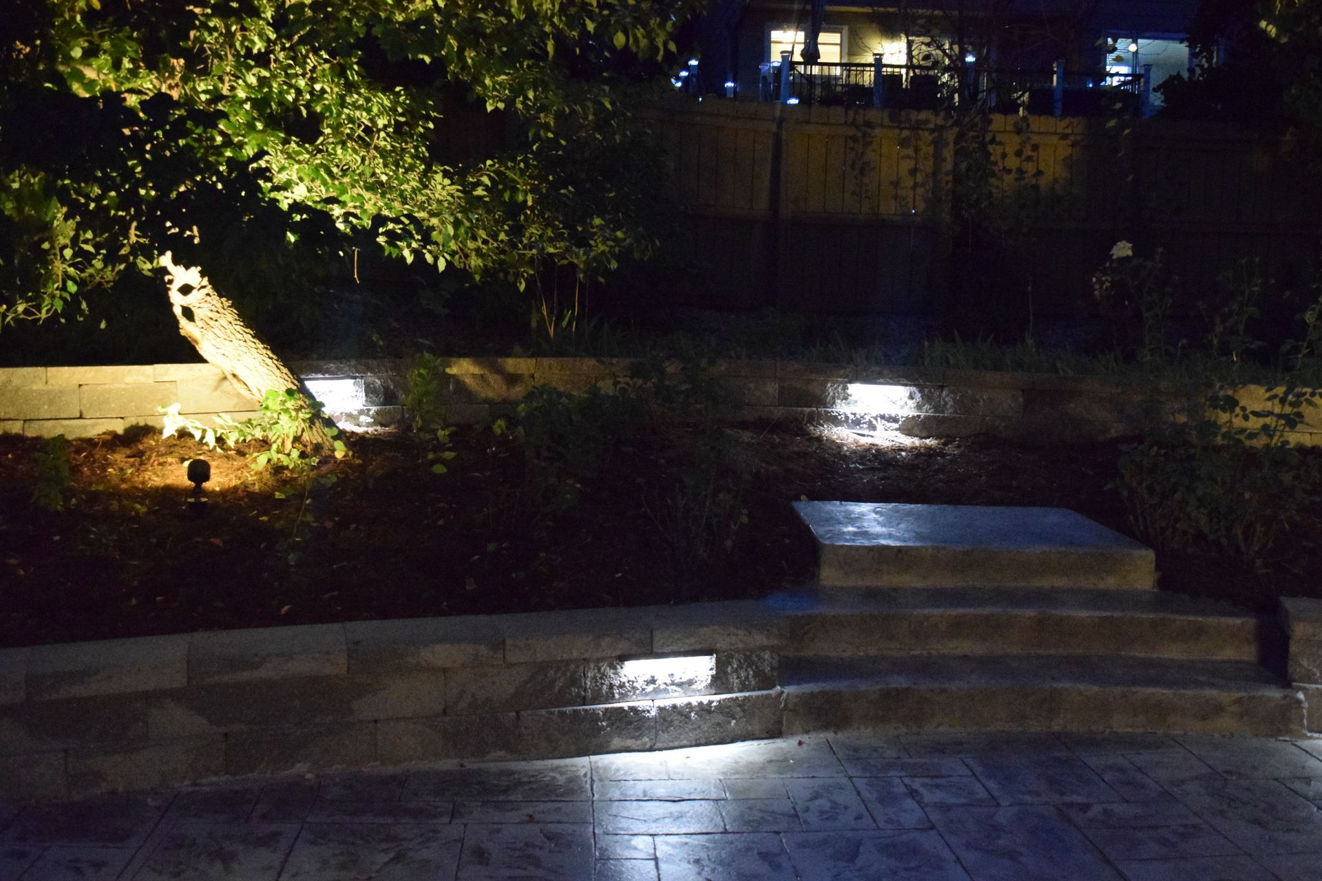 A nighttime landscape with illuminated steps, bushes, and a tree, creating a softly lit garden scene.