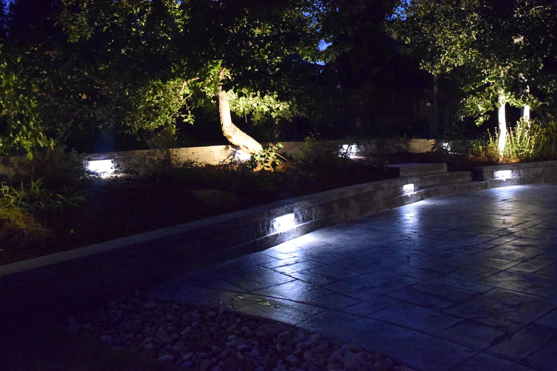 Outdoor scene at night, lit by landscape lights. Trees, brick walls, and a dark paved patio.