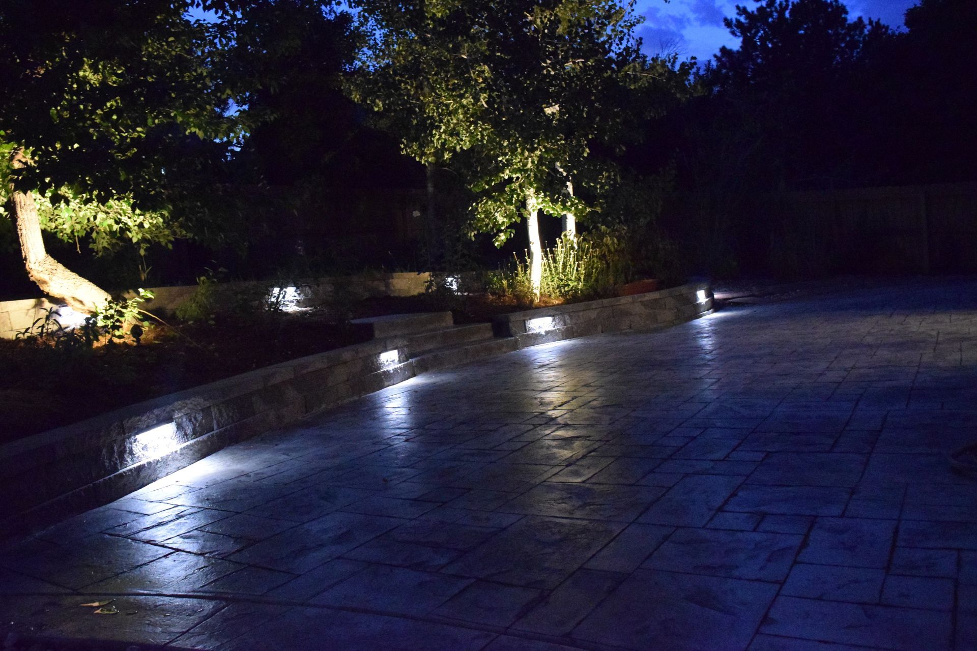 Nighttime view of a lit patio with lights along a stone wall and illuminating trees.