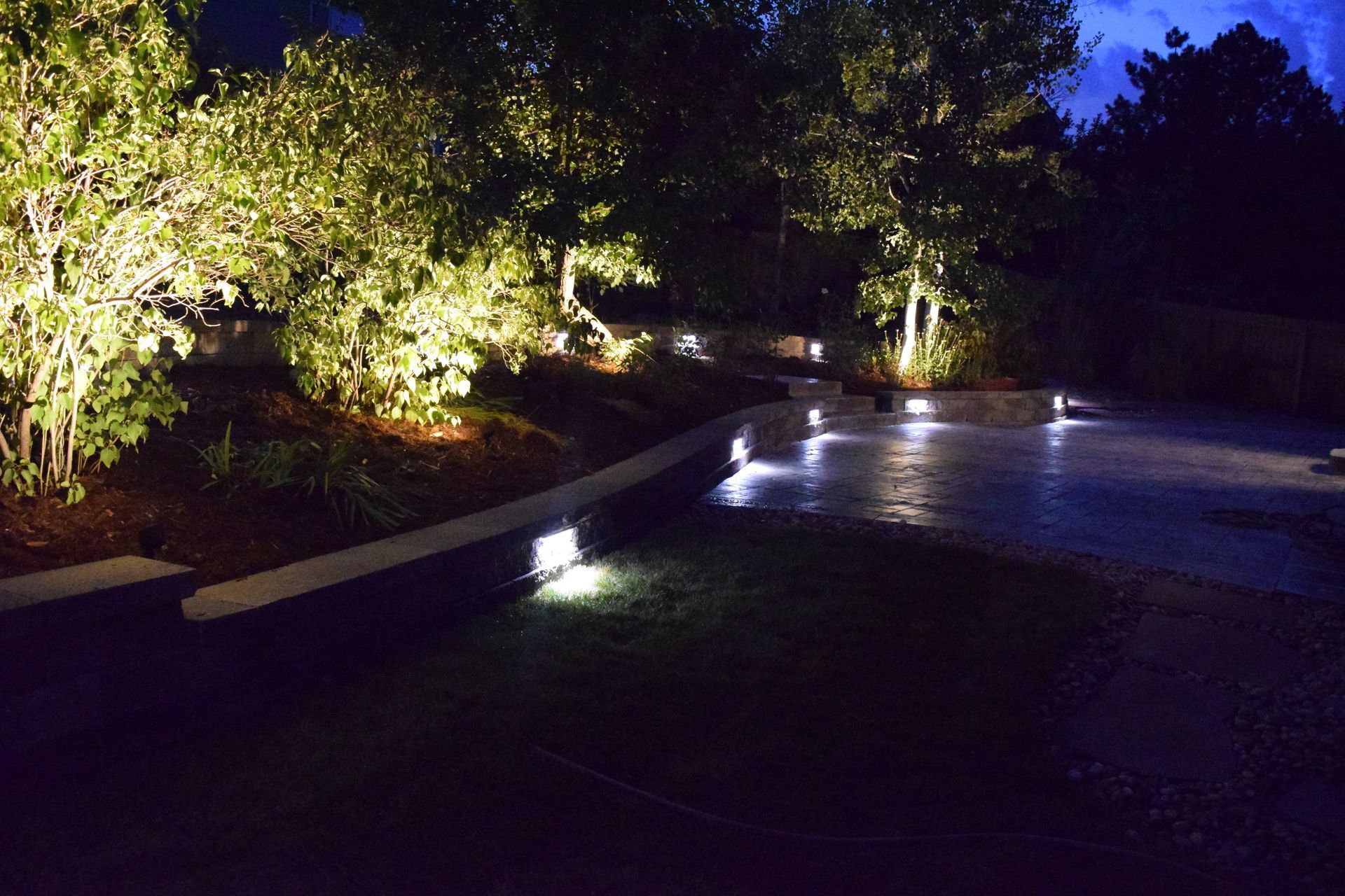 Night view of a yard with landscape lighting illuminating trees, a retaining wall, and a patio.