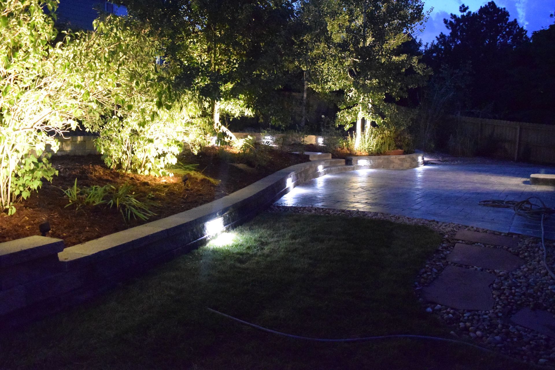 A nighttime shot of a landscaped backyard with trees and a patio illuminated by outdoor lights.