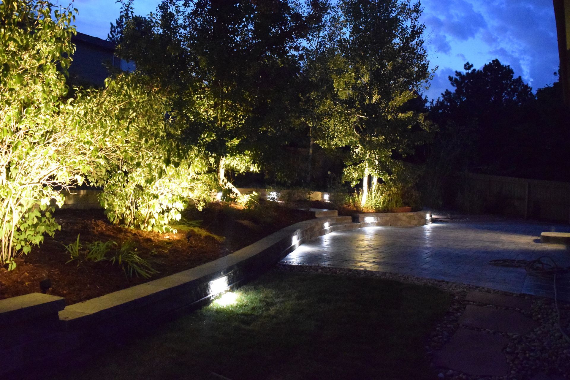 Lit landscaping at night: trees and bushes illuminated, steps with pathway lights, and a stone patio.