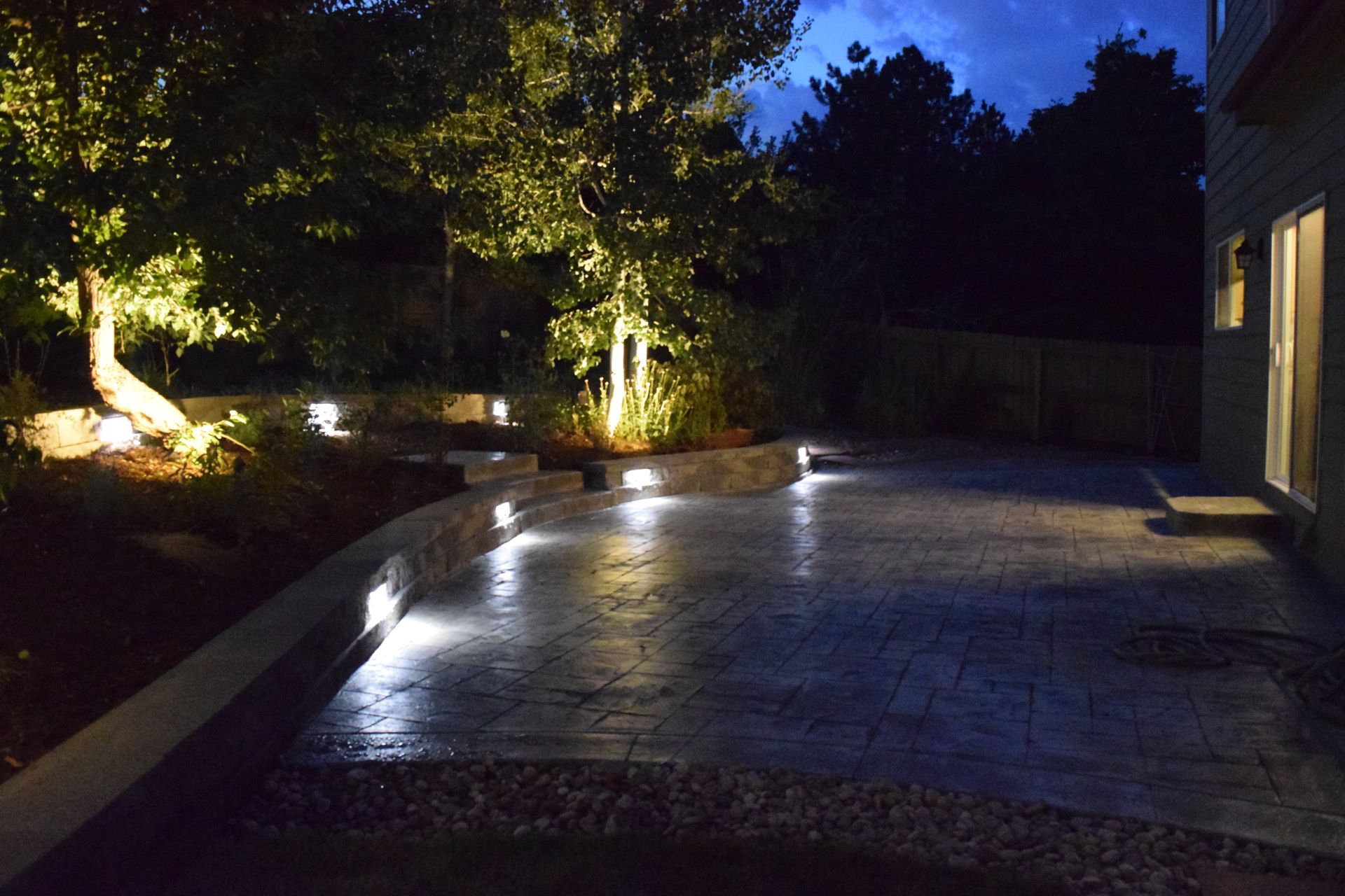 Nighttime backyard patio with up-lit trees, inset lights in retaining walls, and a view into a house.