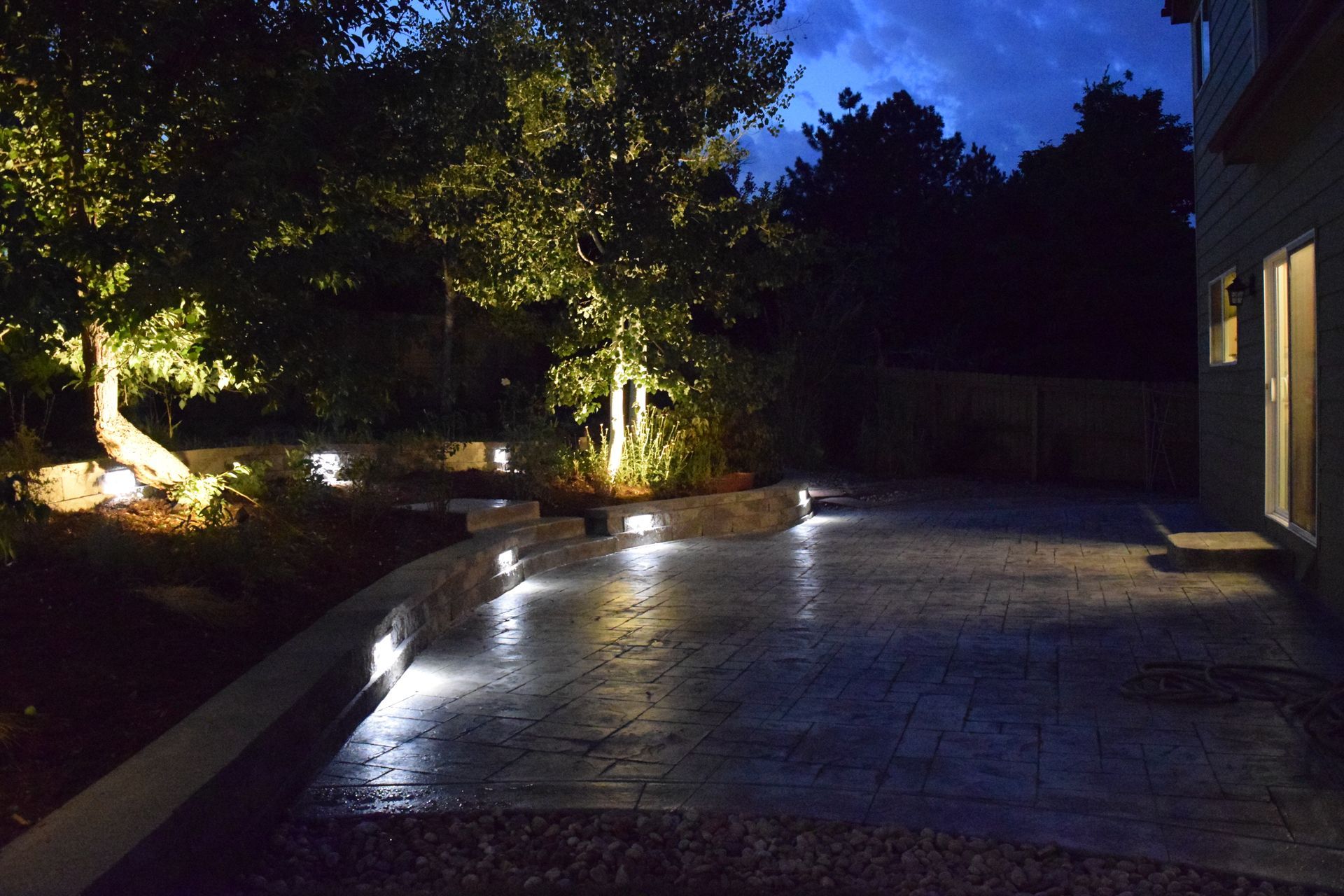 Nighttime view of a patio with built-in lights and illuminated trees, next to a house.