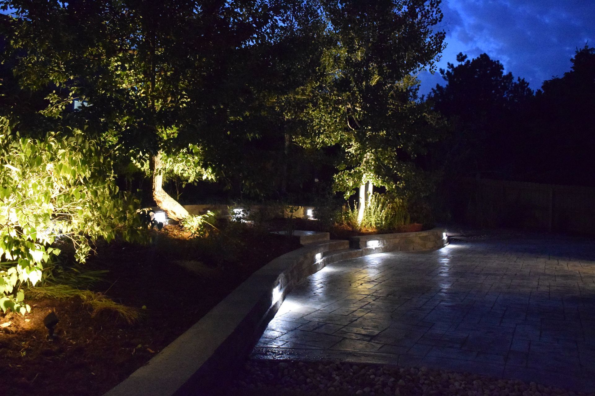 Night landscape with illuminated trees, pathway, and retaining wall; blue sky.