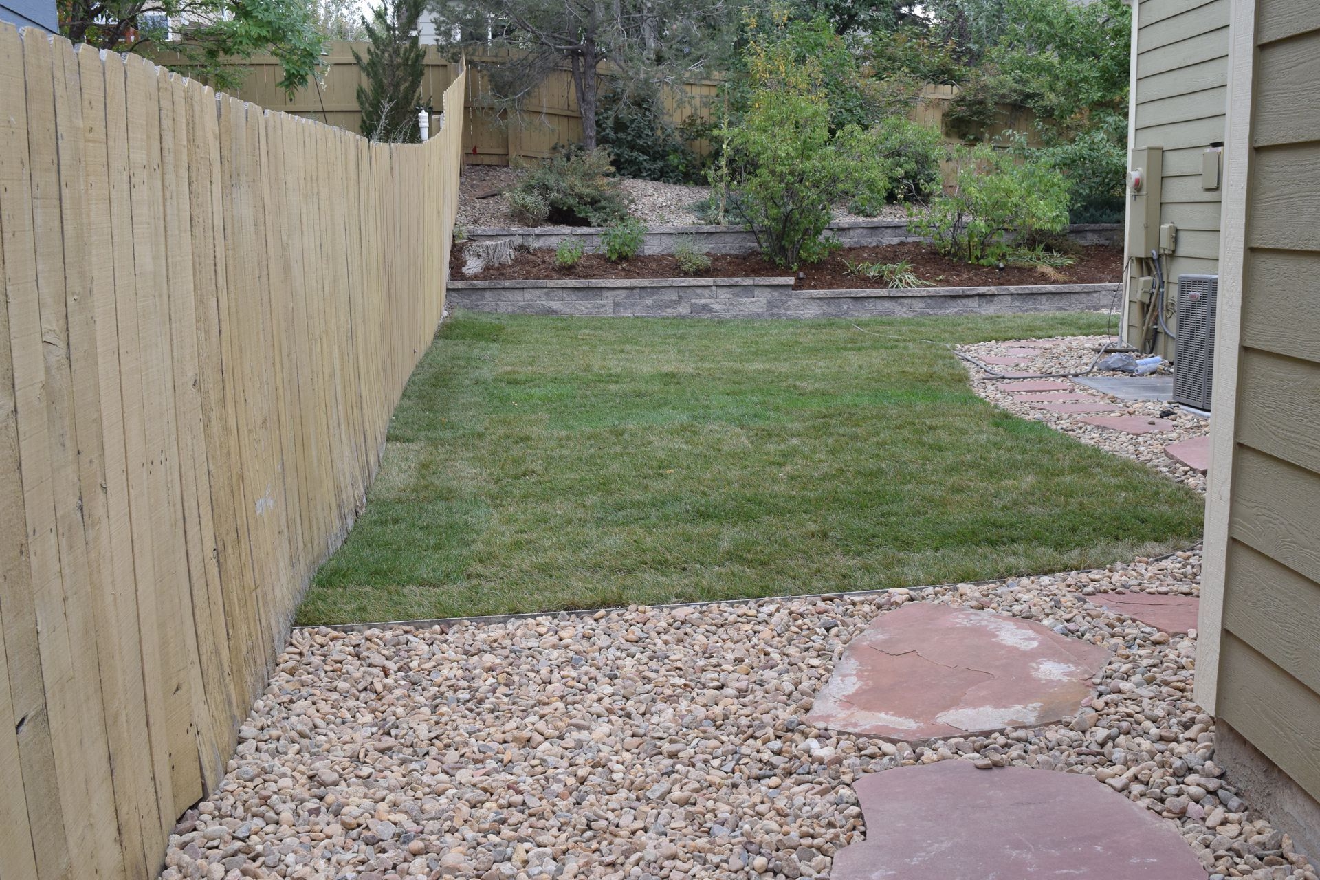 A backyard with a wooden fence, lawn, gravel, and flagstone path.