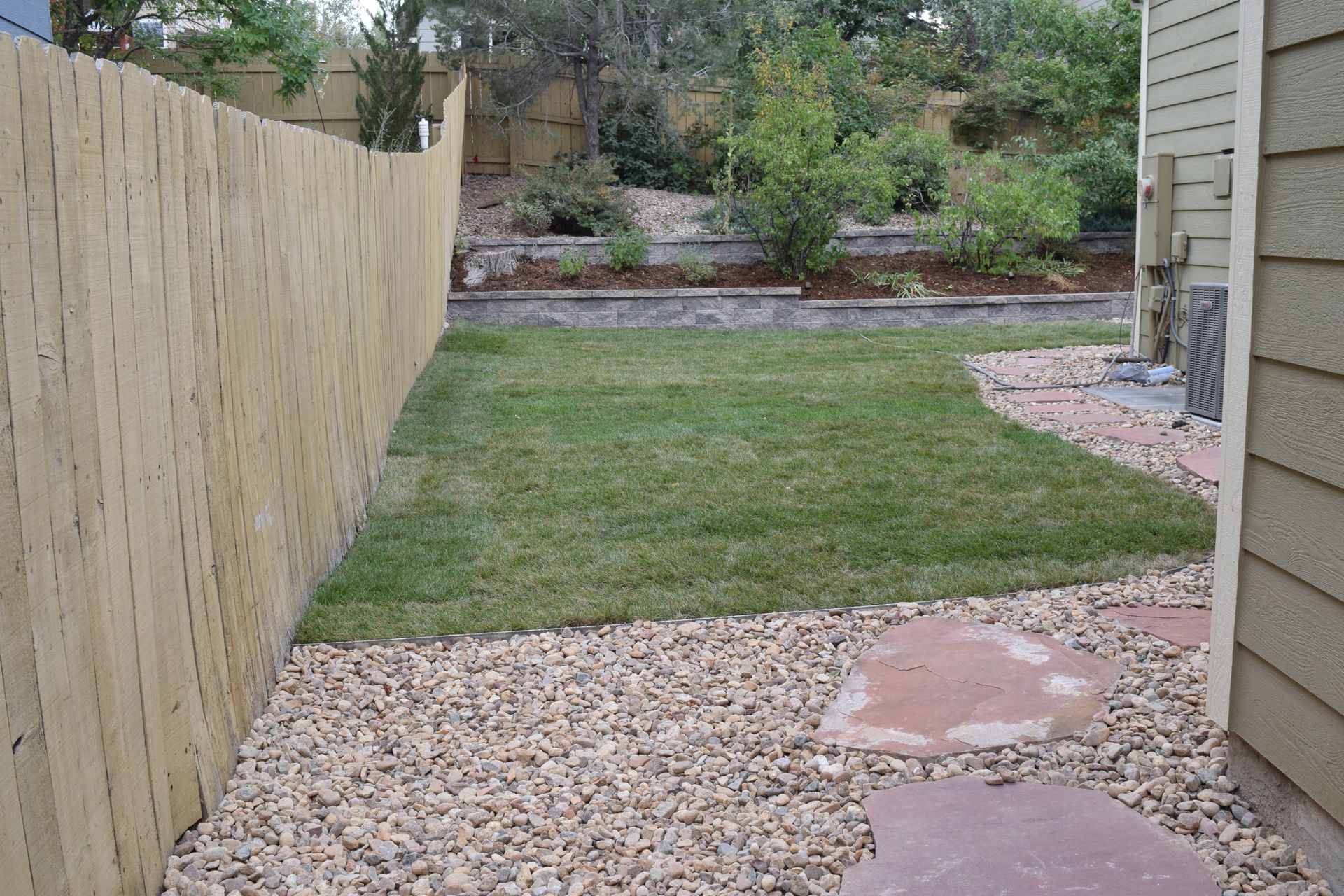 A backyard with a wooden fence, green lawn, stone path, and gravel.