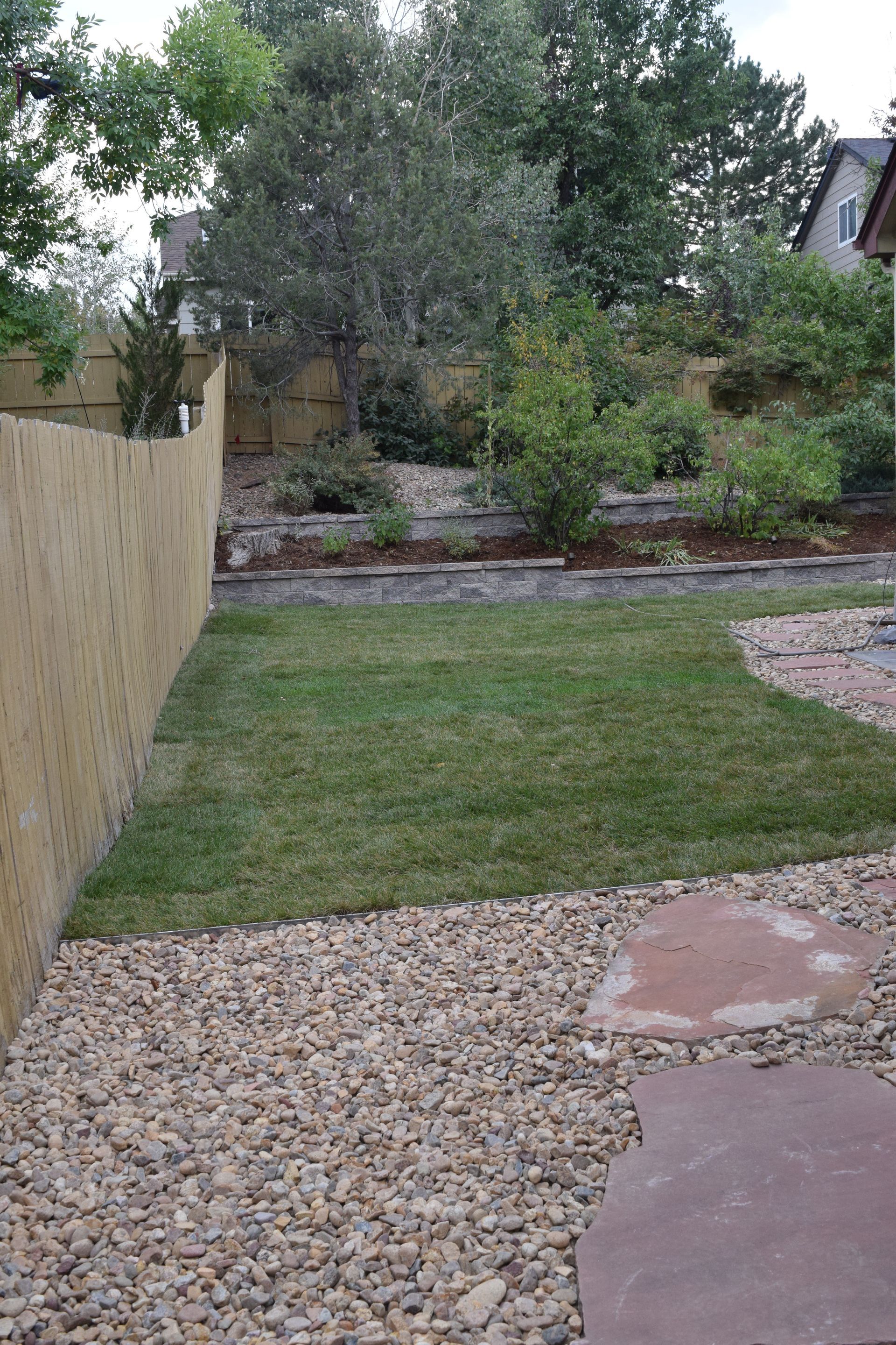 A backyard with a wooden fence, grass, gravel path, and tiered garden beds.