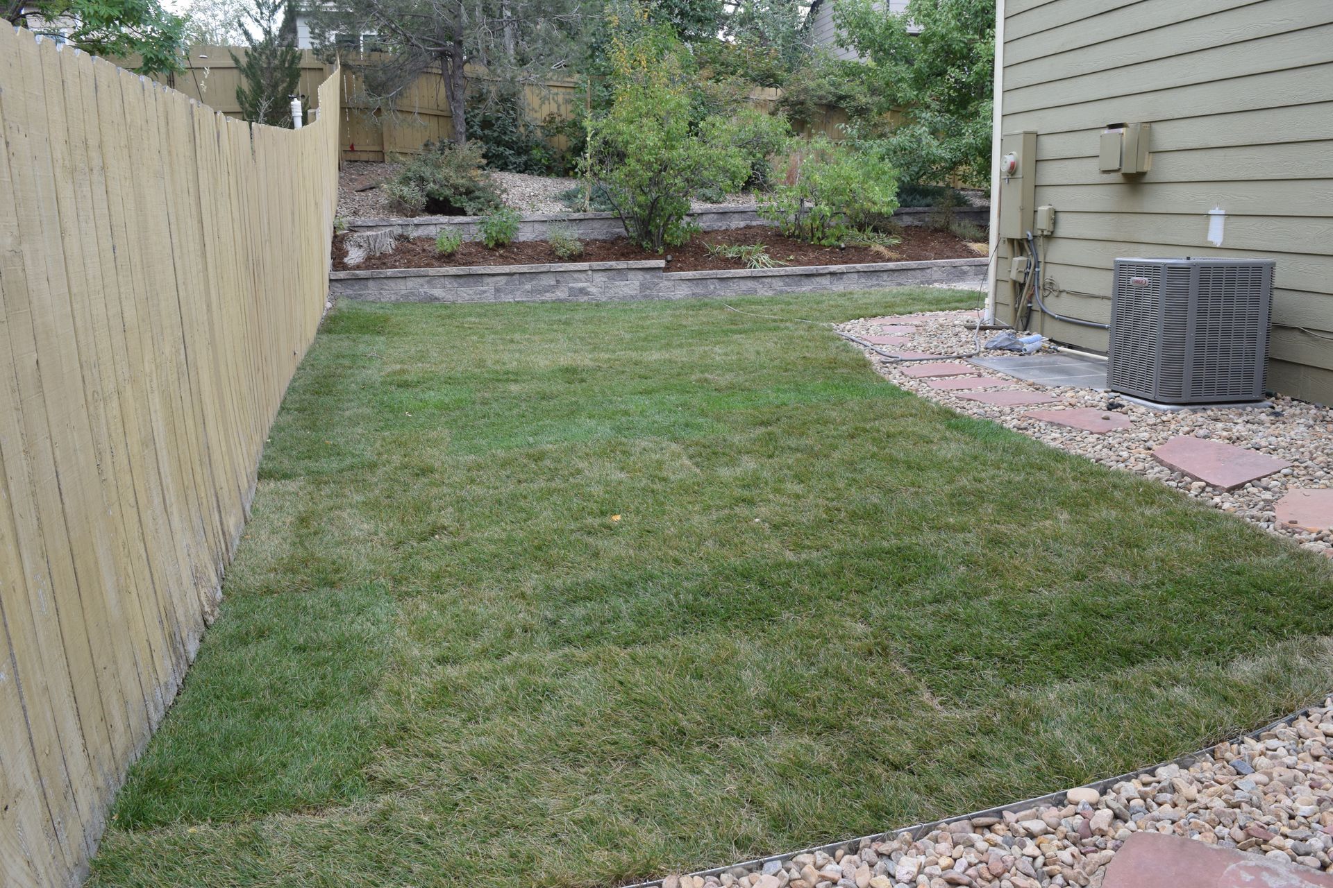 A grassy backyard bordered by a wooden fence, rock path, and a building with a gray air conditioning unit.