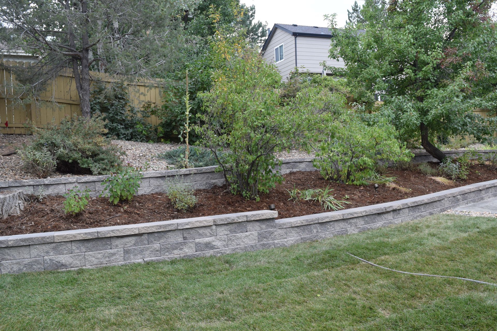 Stone retaining walls with plants and mulch in a backyard, trees, and a house in the background.