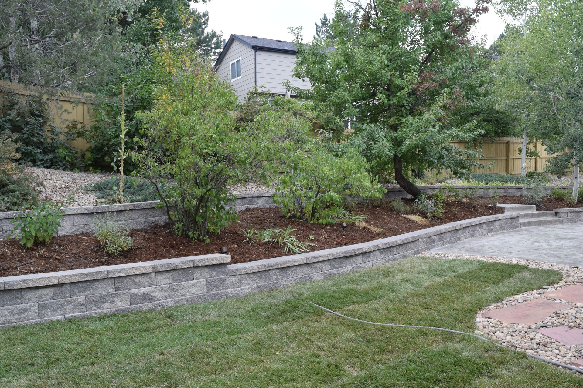 Backyard with tiered landscaping and retaining walls, trees, grass, and a paved area.