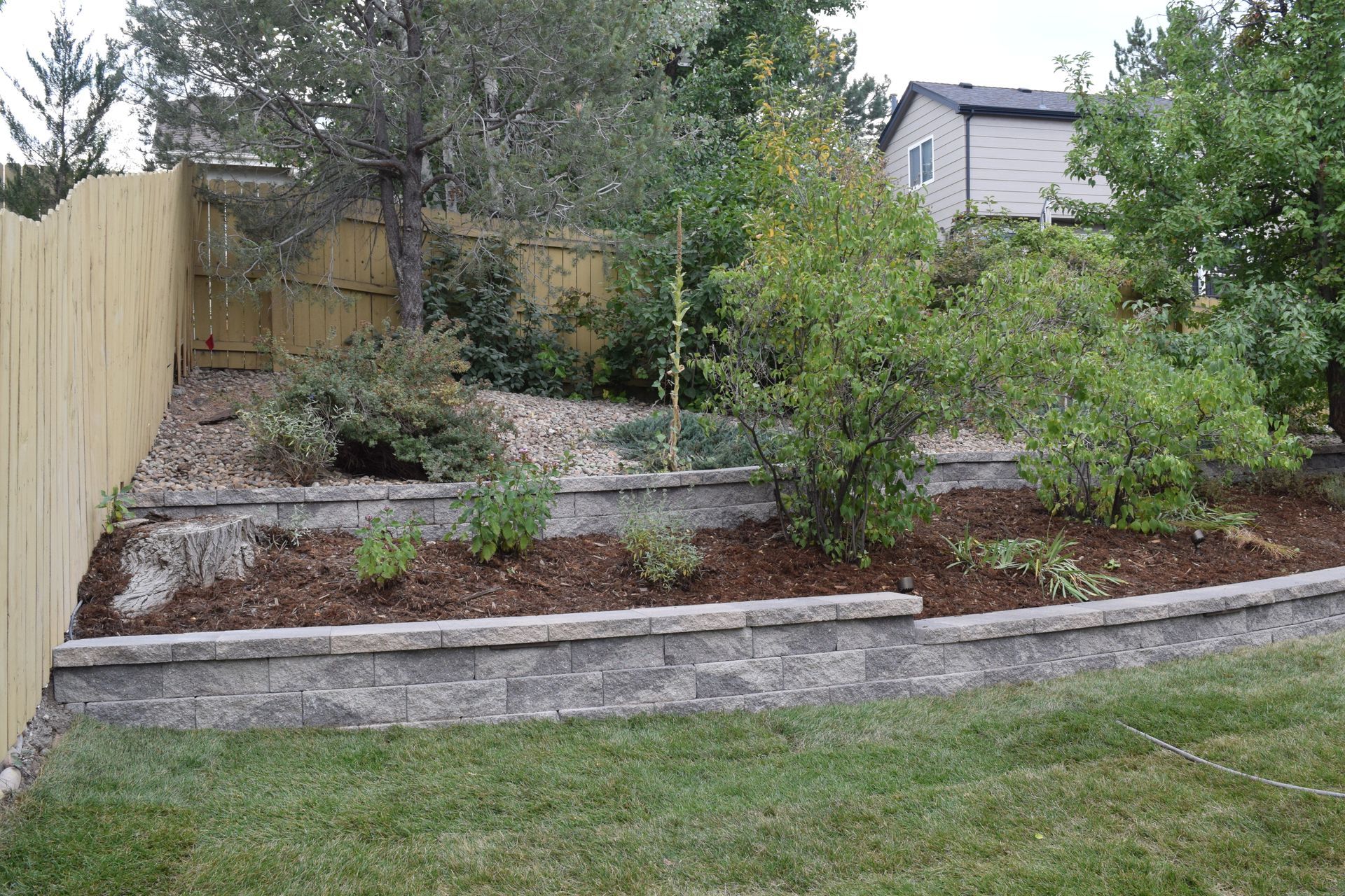 Backyard with tiered stone retaining walls, plants, mulch, wooden fence, and grass.
