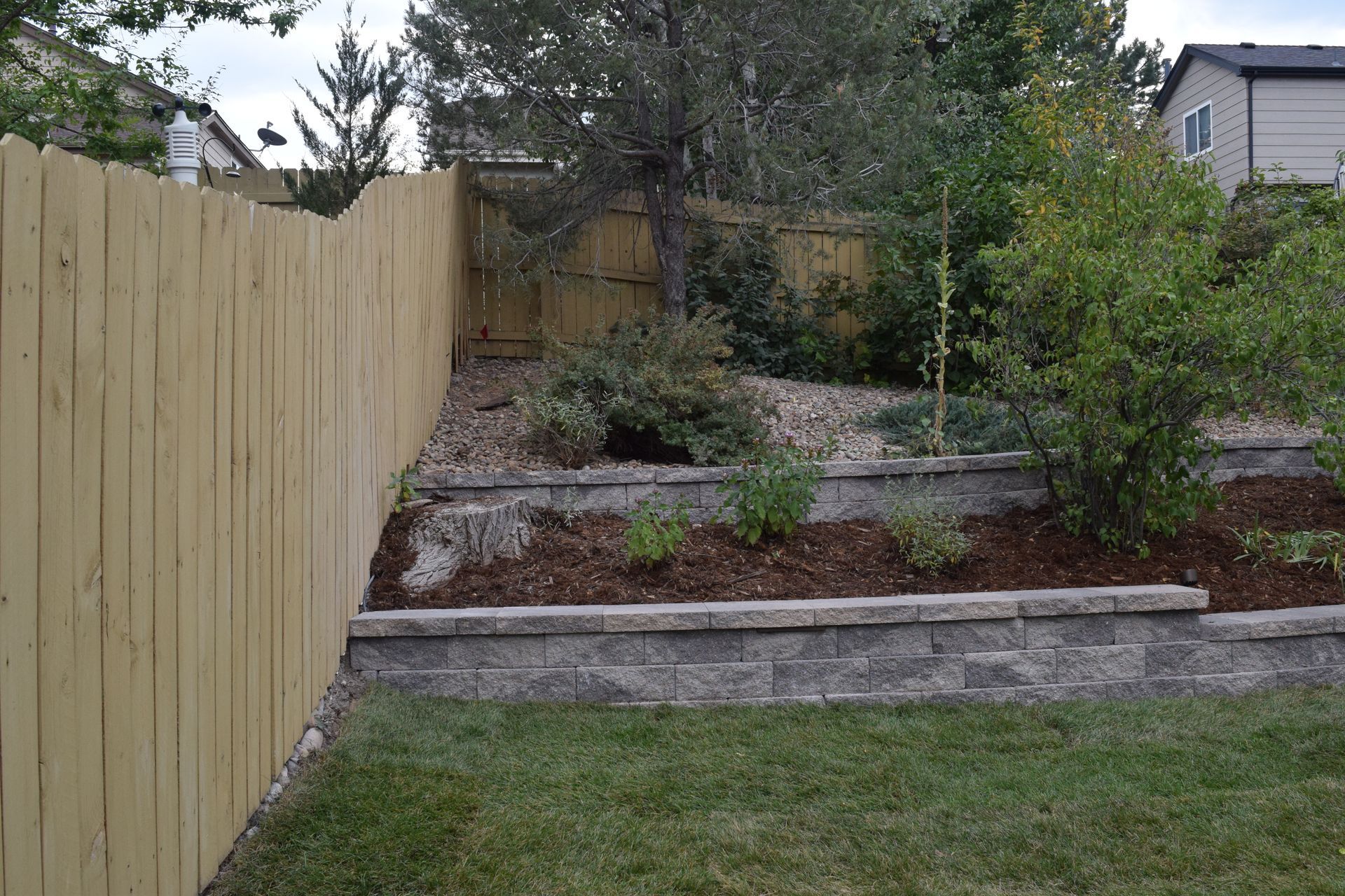 Wooden fence bordering a backyard with tiered landscaping, retaining walls, and greenery.