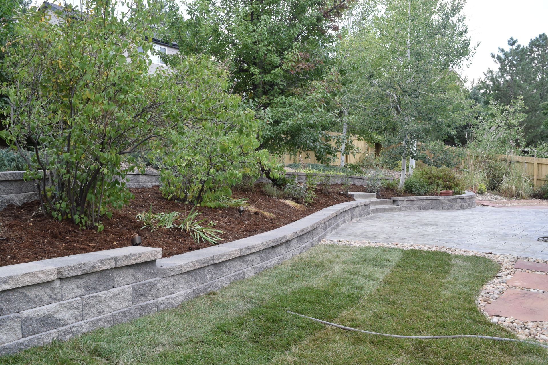 Stone retaining wall with greenery and mulch bordering a patio and lawn.
