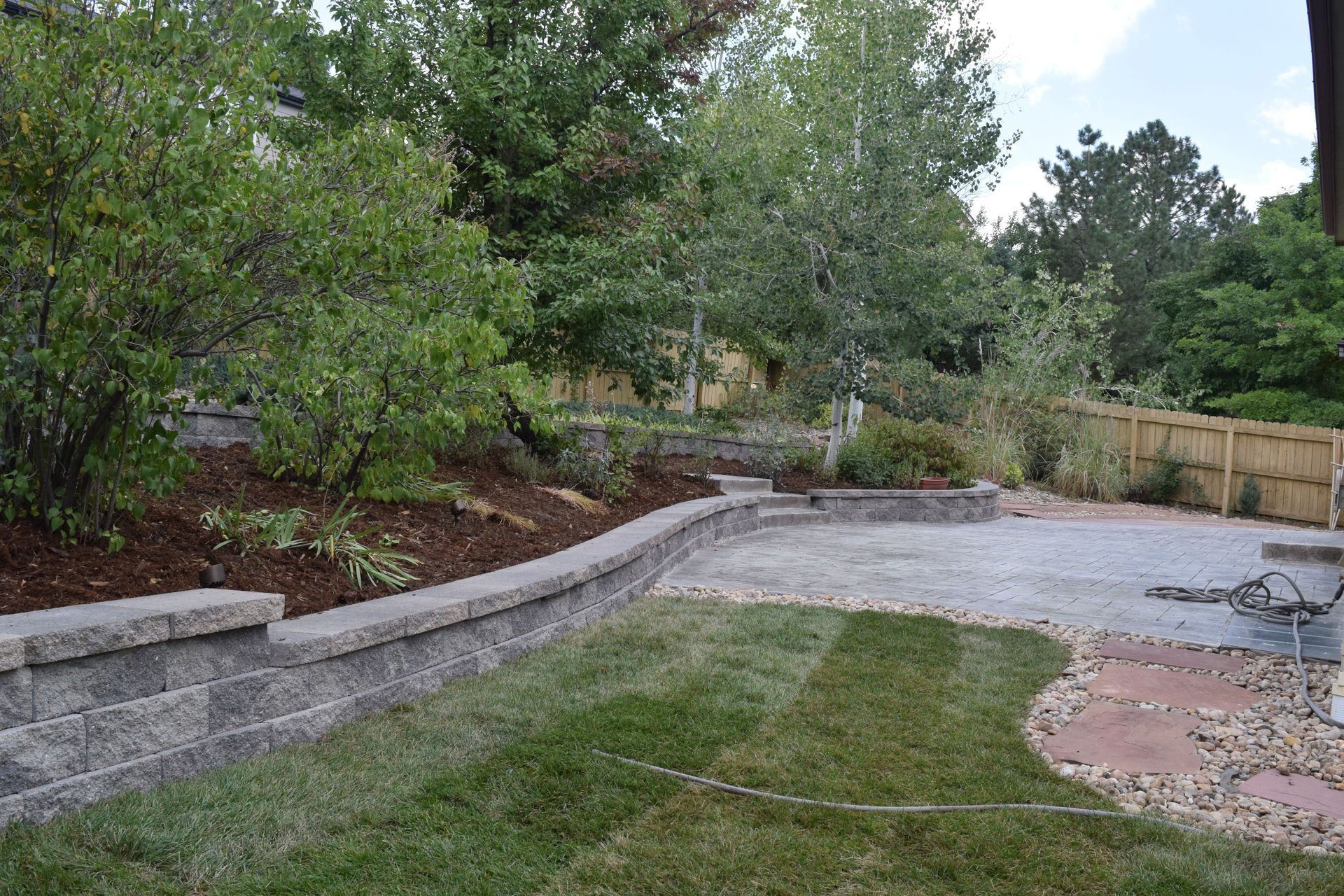 Backyard with a retaining wall, gravel patio, green lawn, and trees.