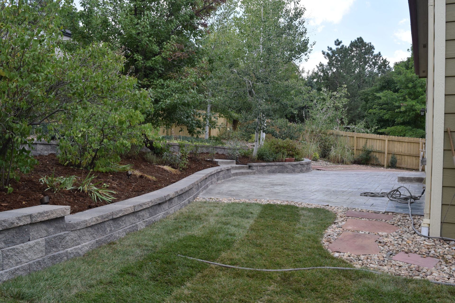 A backyard with a patio, retaining wall, lawn, and trees.