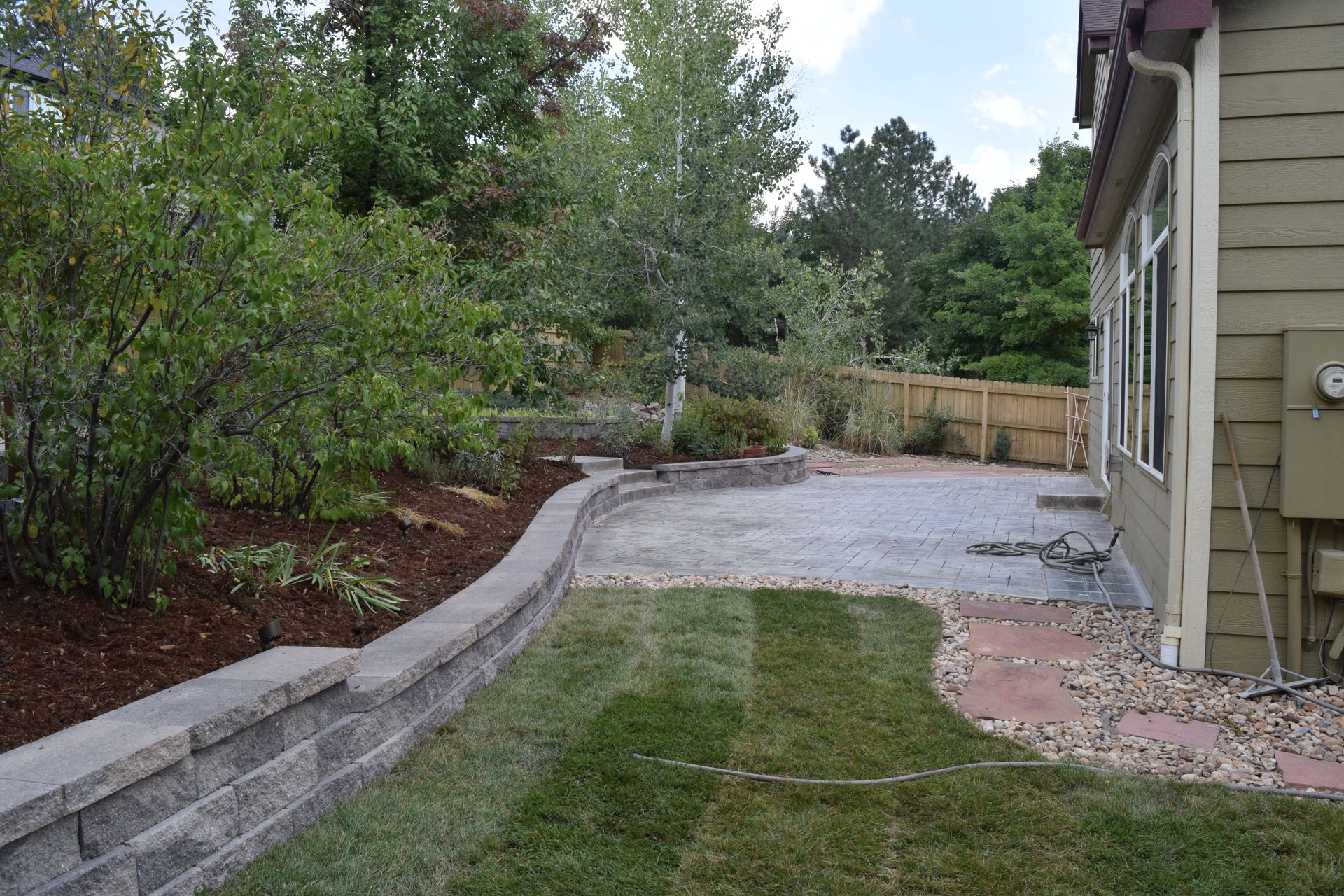 Backyard with retaining wall, patio, lawn, and trees. House on right.