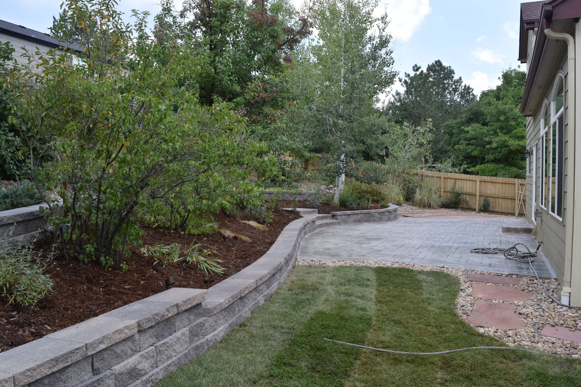 Backyard with retaining walls, patio, grass, and trees. Brown mulch and brick walls, green lawn.