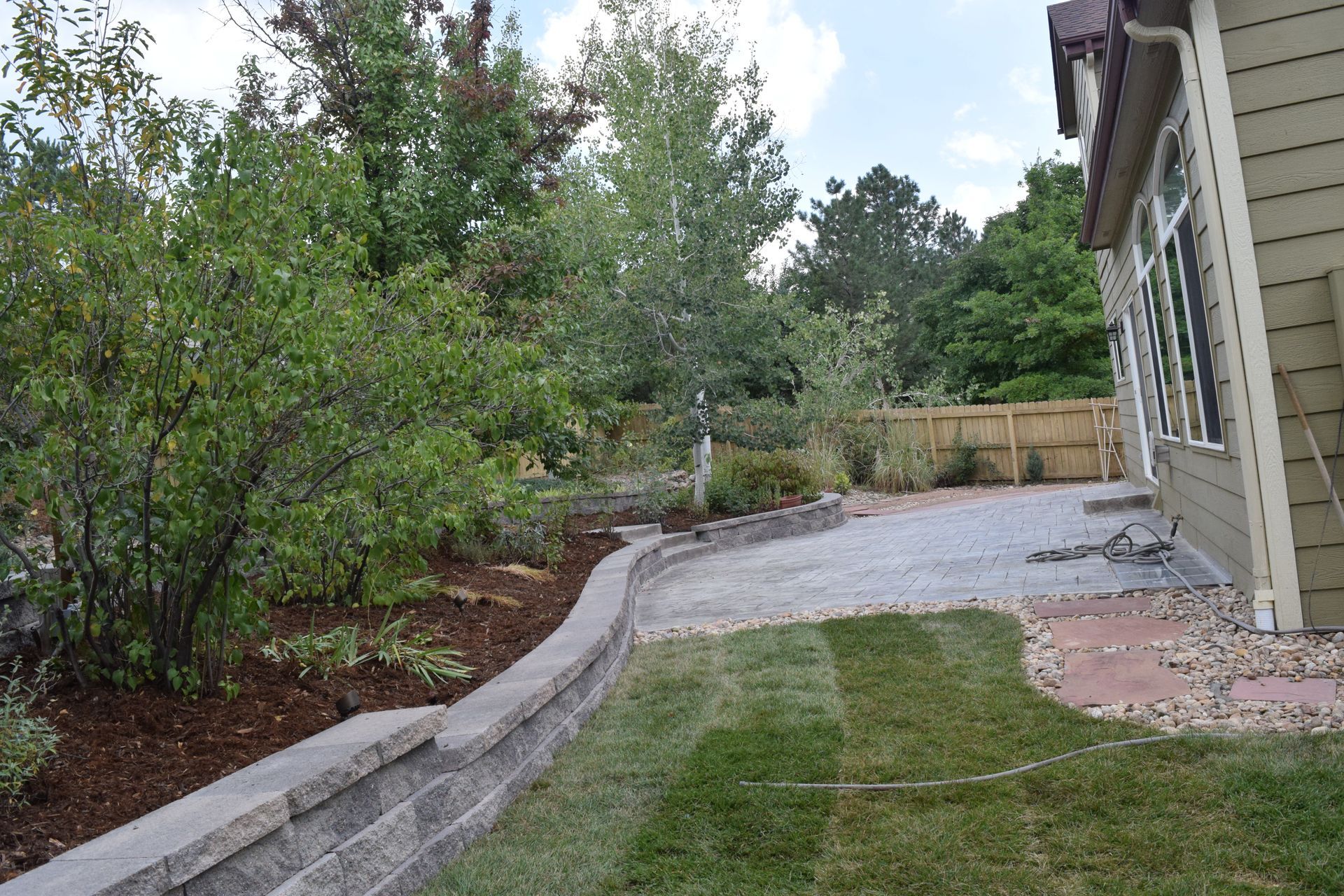 A backyard with a retaining wall, gravel patio, green grass, trees, and a house.