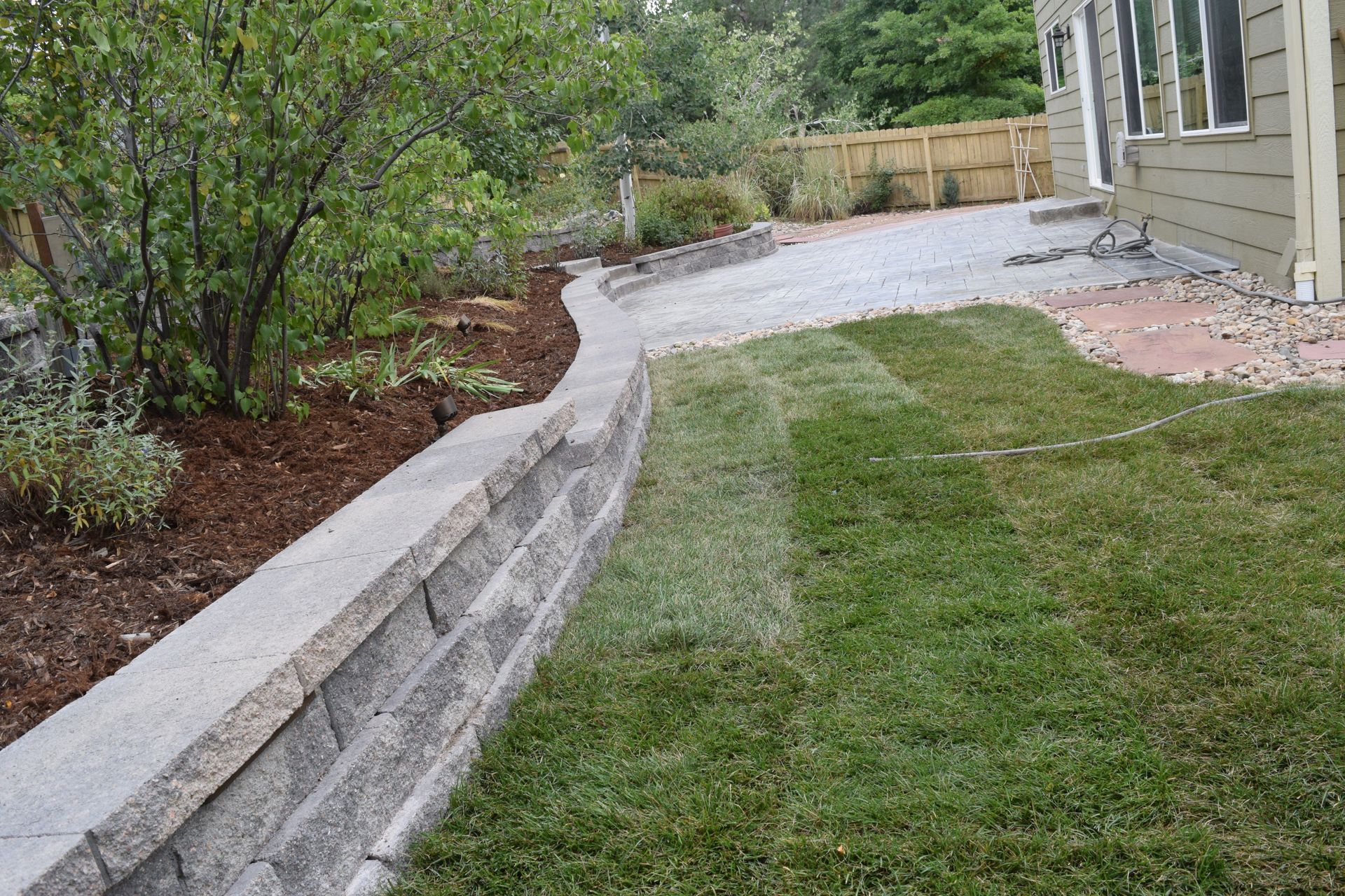 A retaining wall separates a grassy lawn and mulched area, beside a driveway and house.