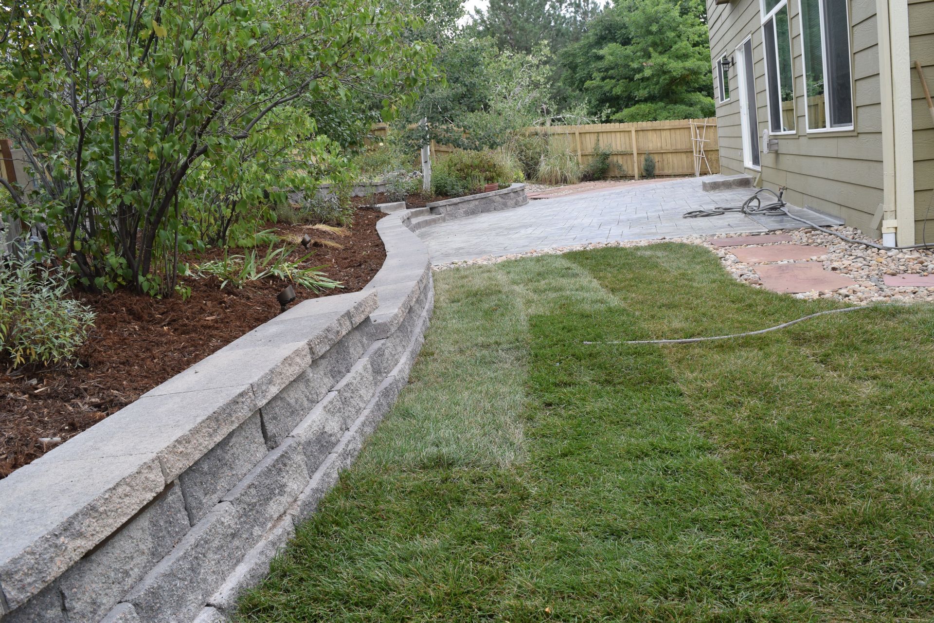 Stone retaining wall borders a lawn, separating it from a gravel patio and landscaping.