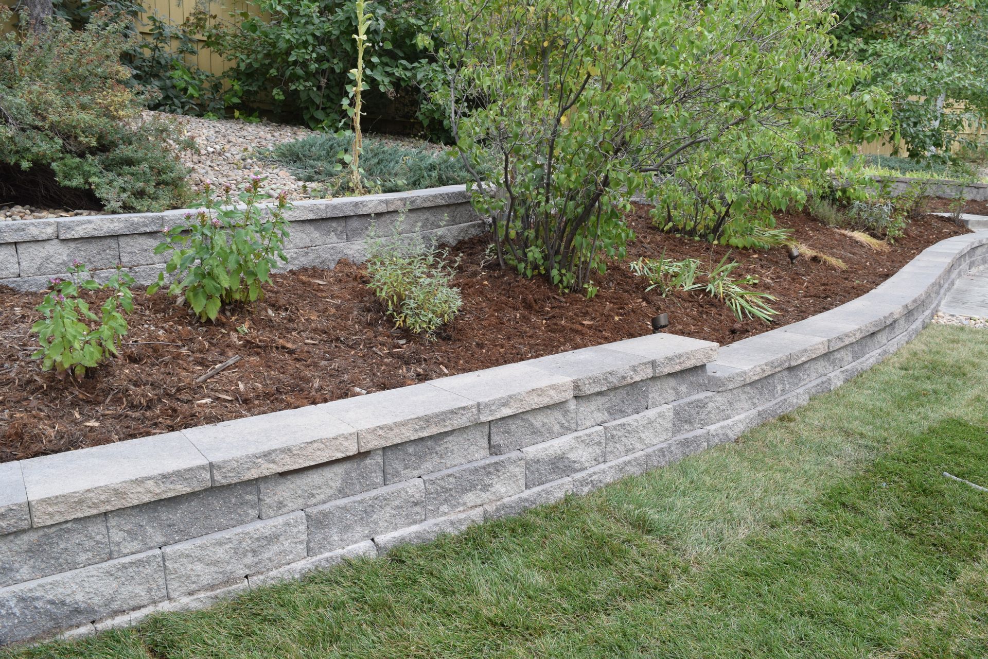Retaining wall with tiered garden beds and mulch, with grass in the foreground and plants.