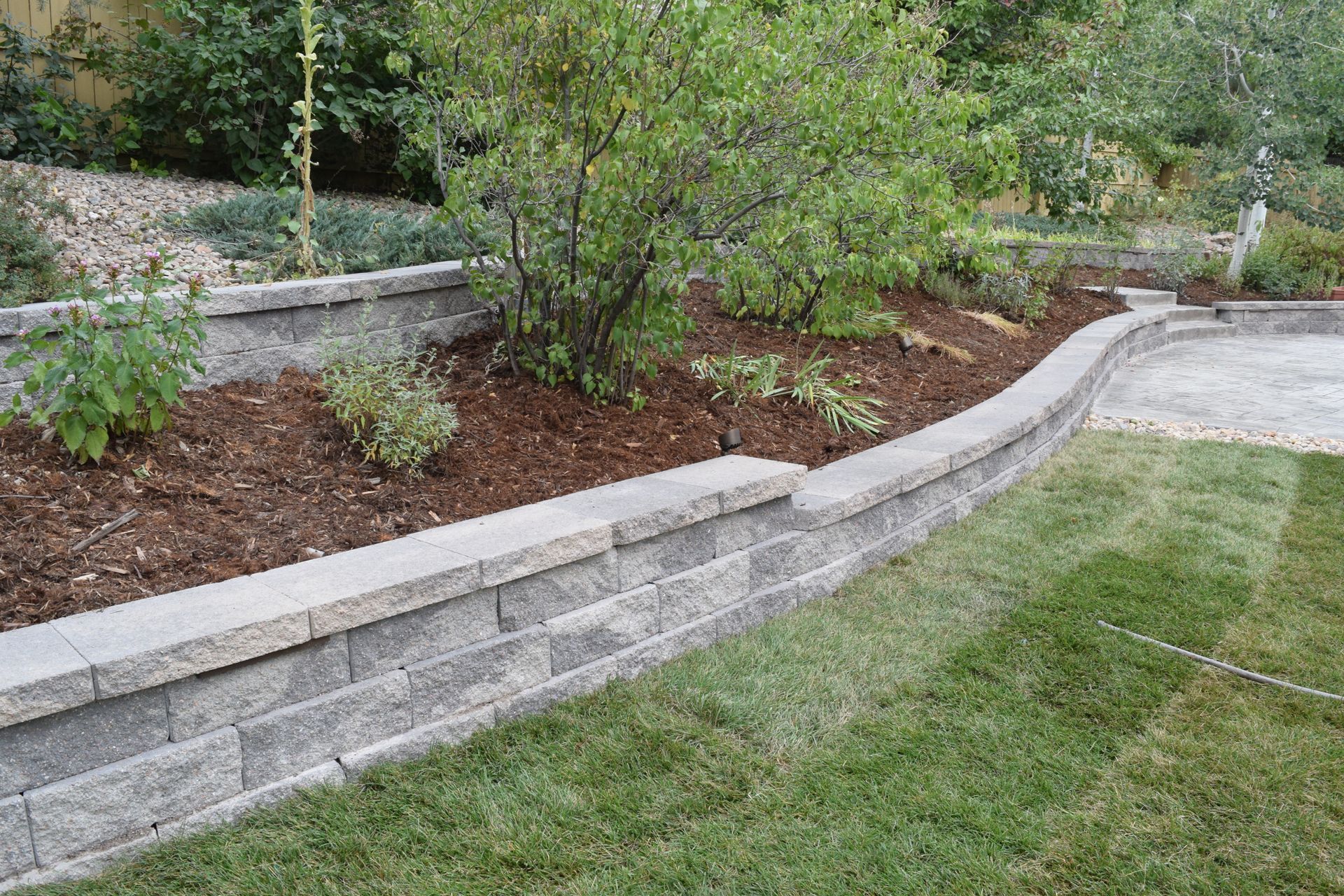 Retaining wall of gray blocks borders a landscaped bed with mulch and plants, alongside a green lawn.