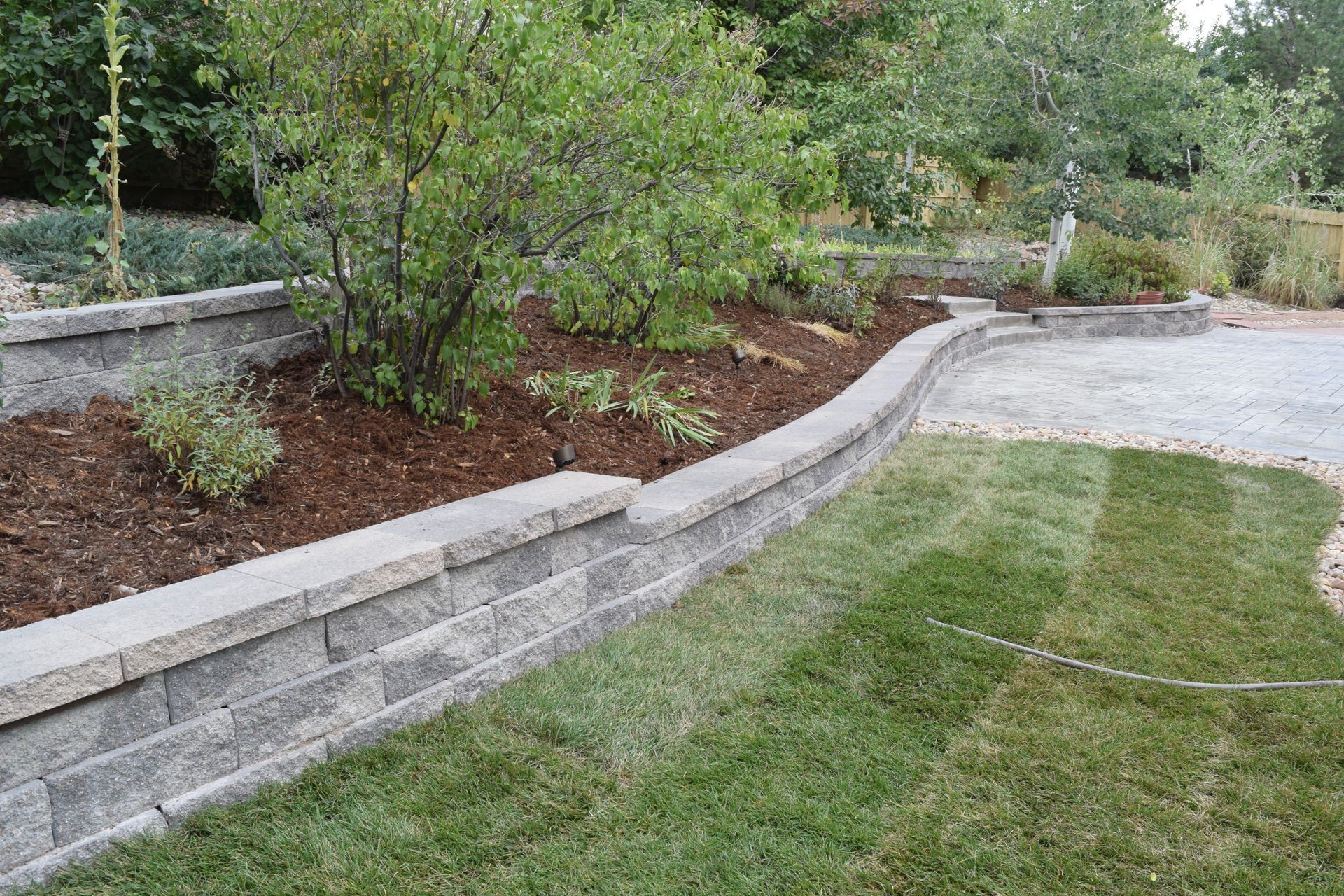 Stone retaining wall bordering a mulched garden bed with shrubs, next to a grass lawn and gravel path.