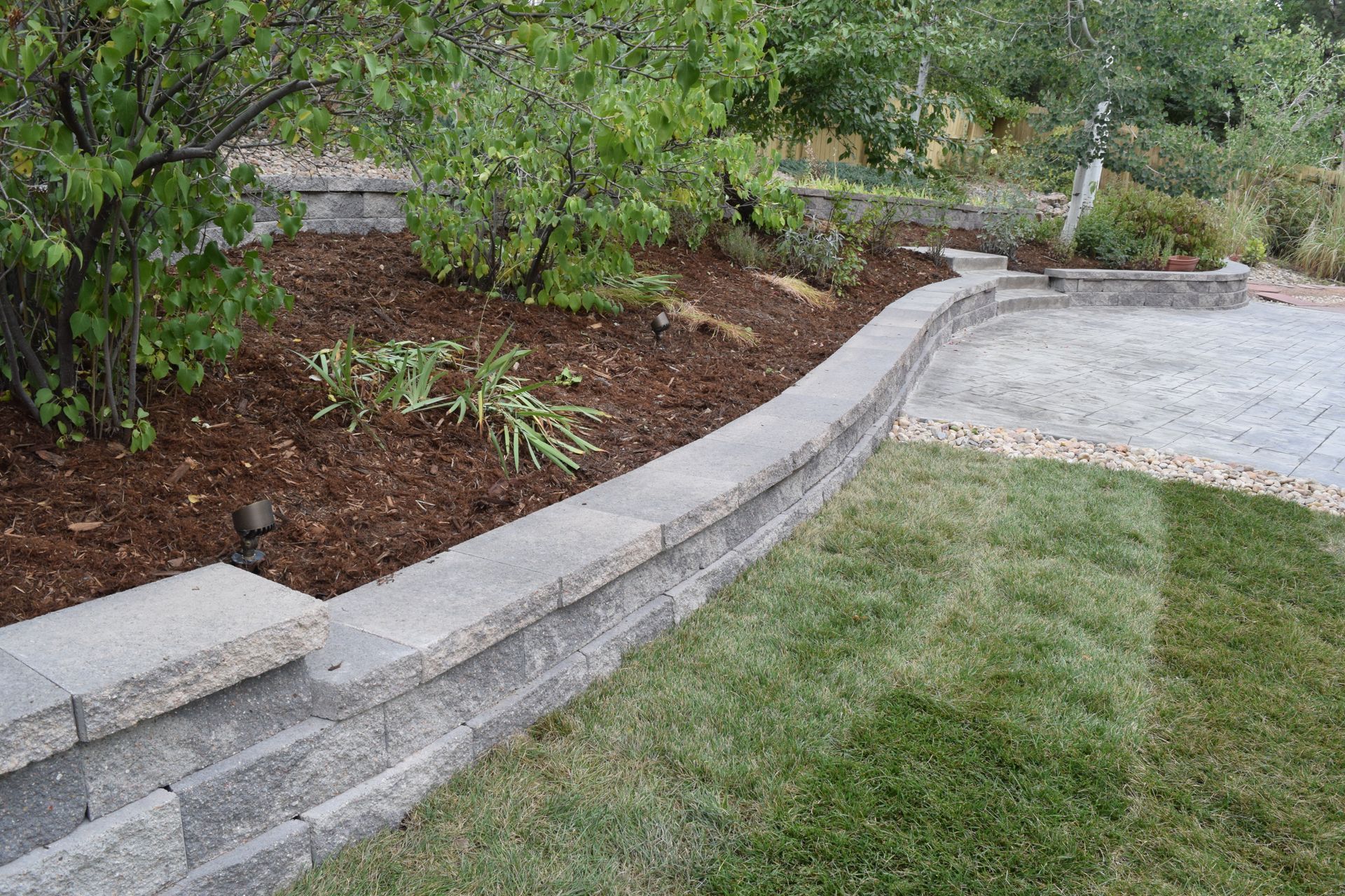 Stone retaining wall with plants, mulch, and lawn. A patio is visible in the background.
