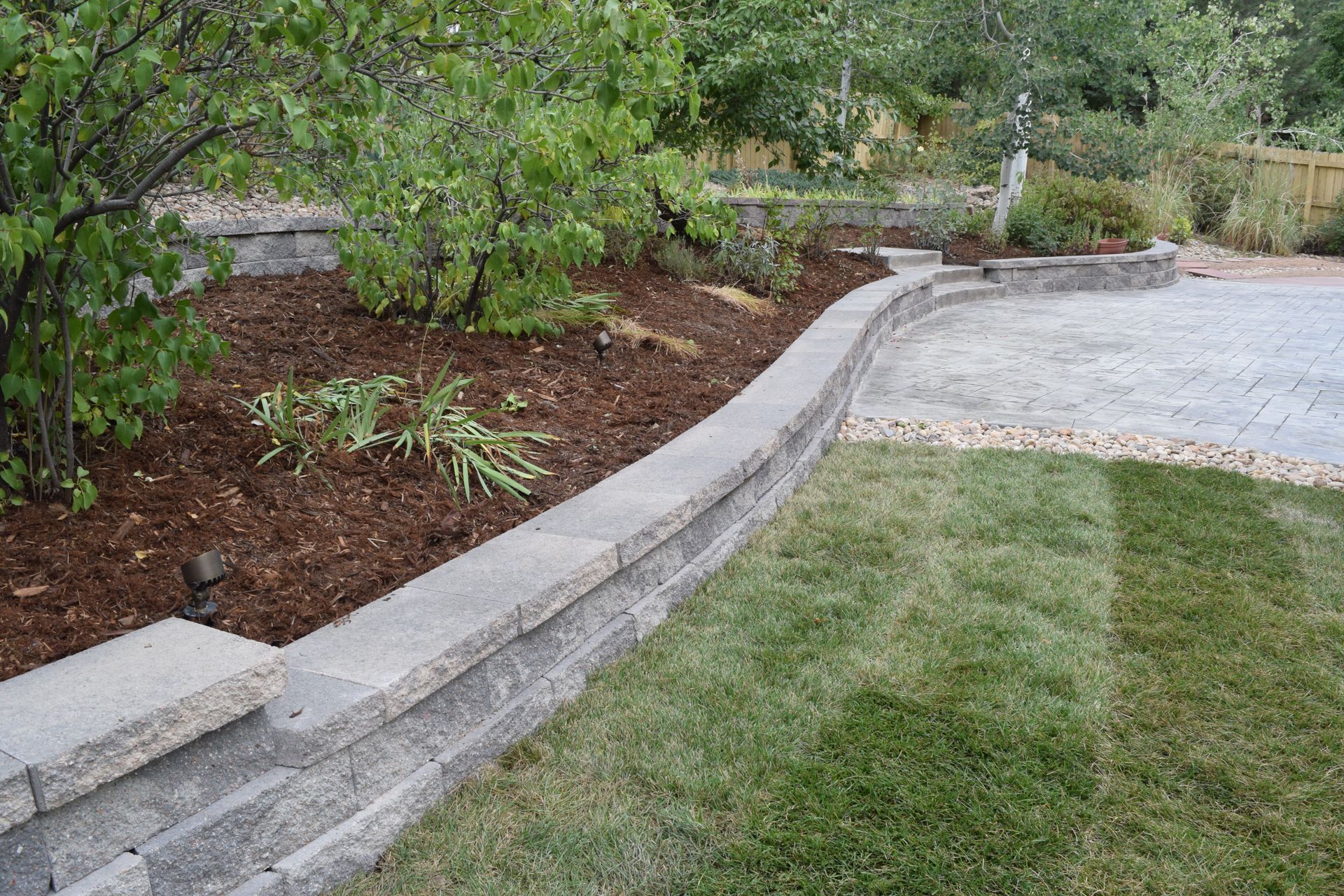 Low retaining wall with mulch, plants, and grass, separating a patio area from a lawn.