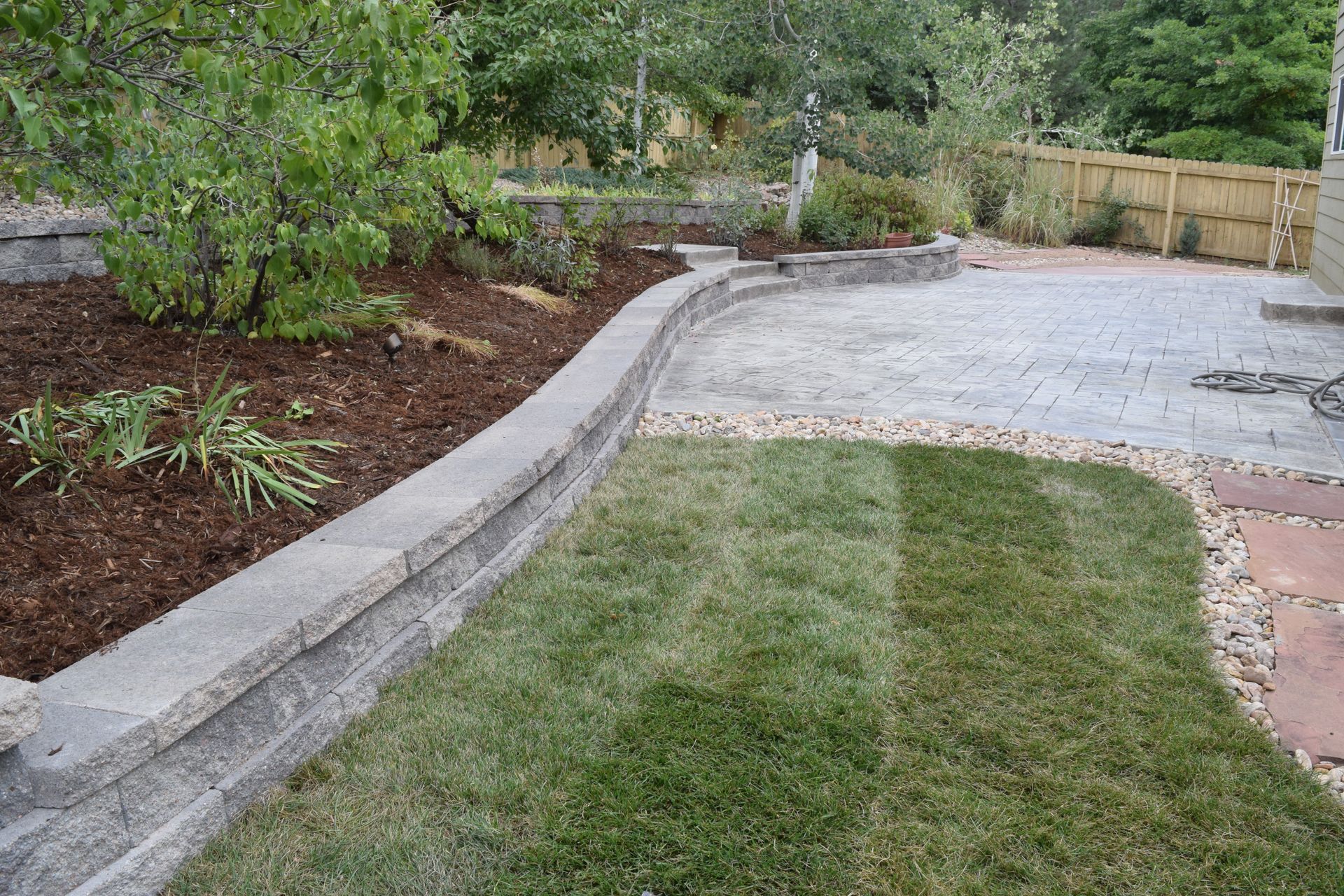 Lawn and stone patio bordered by a low stone wall, mulch, and greenery; a sunny day.