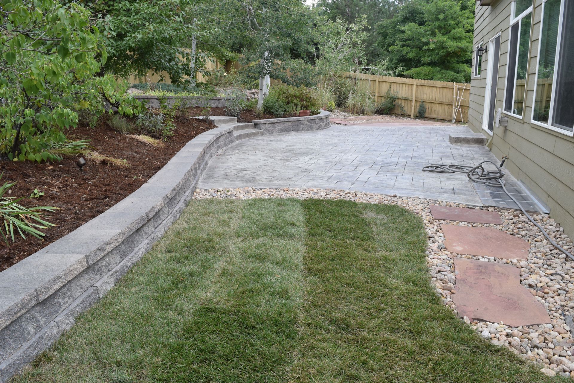 Backyard with lawn, patio, retaining wall, and landscaping; greenery and neutral tones.