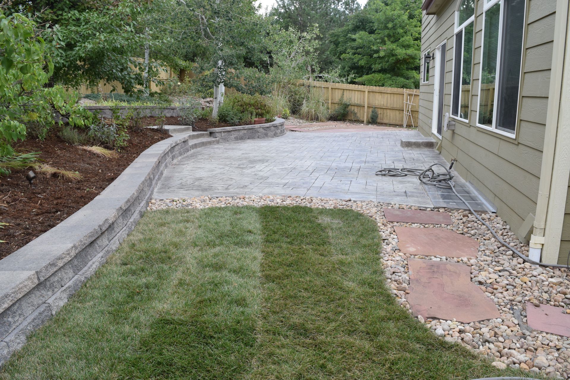 Backyard patio with lawn, stone wall, and landscaping; a house is on the right.
