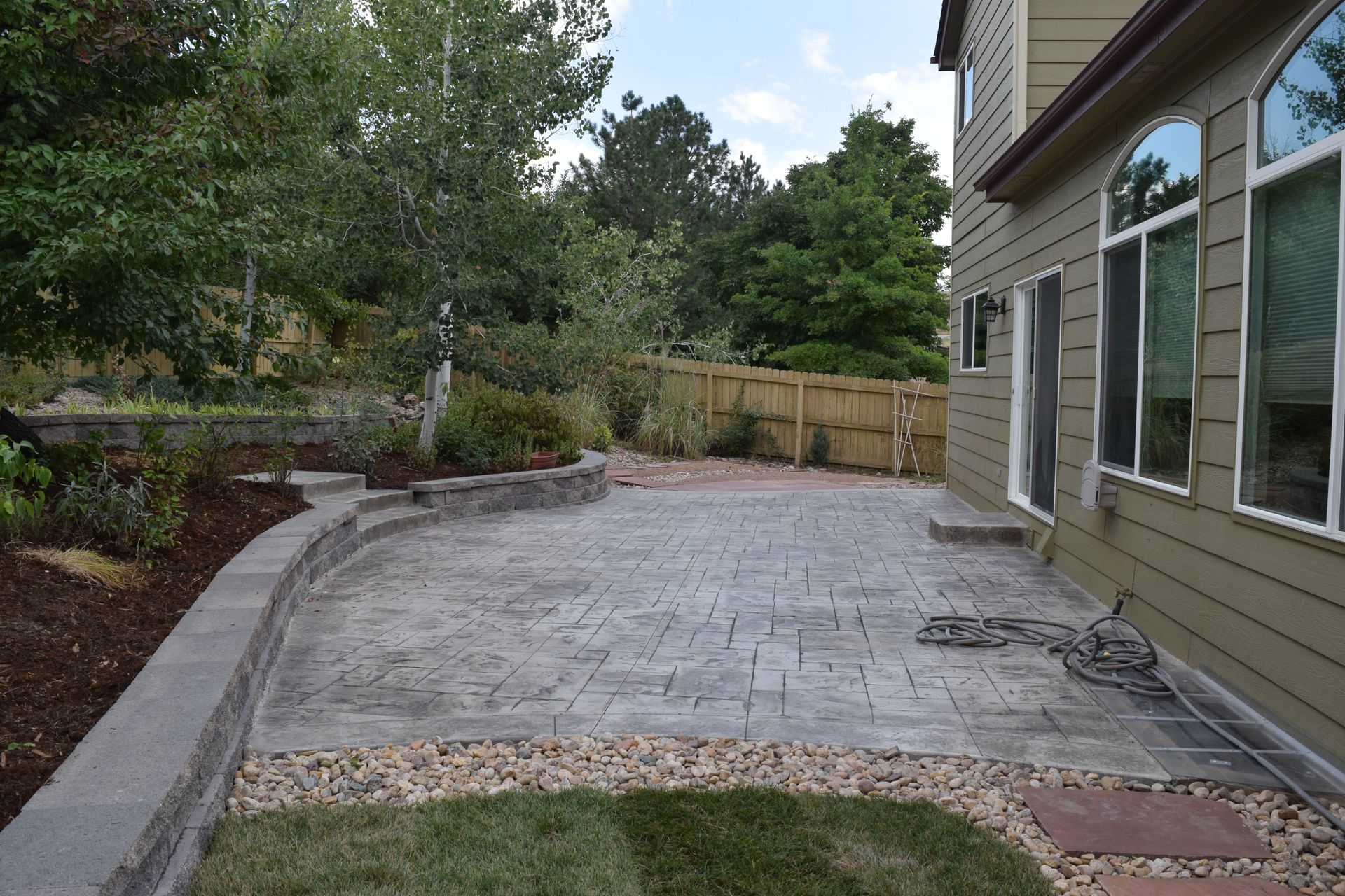 Patio with brick pavers, raised garden bed, house, trees, and wooden fence.
