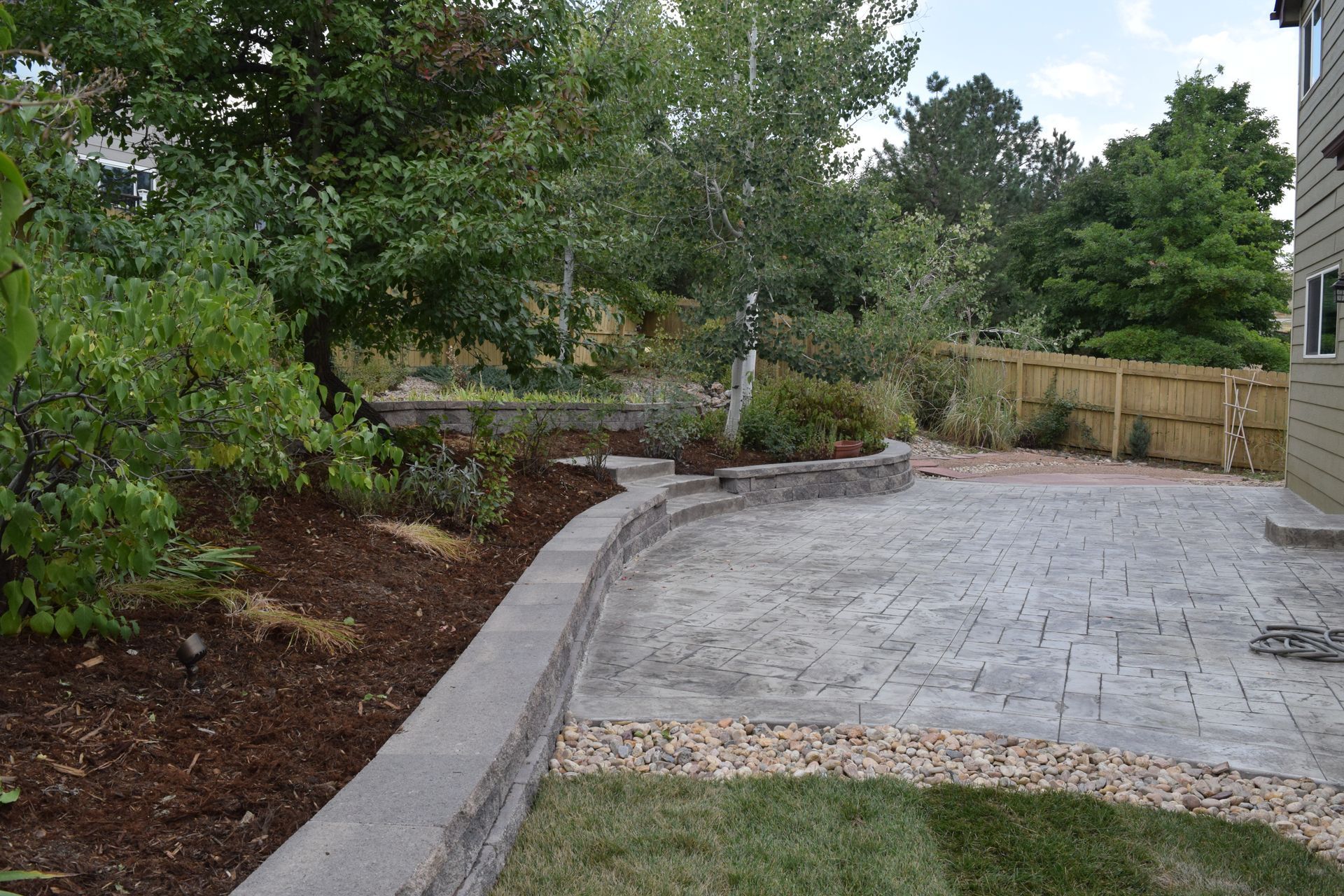 Backyard patio with retaining wall, trees, mulch, and wooden fence.