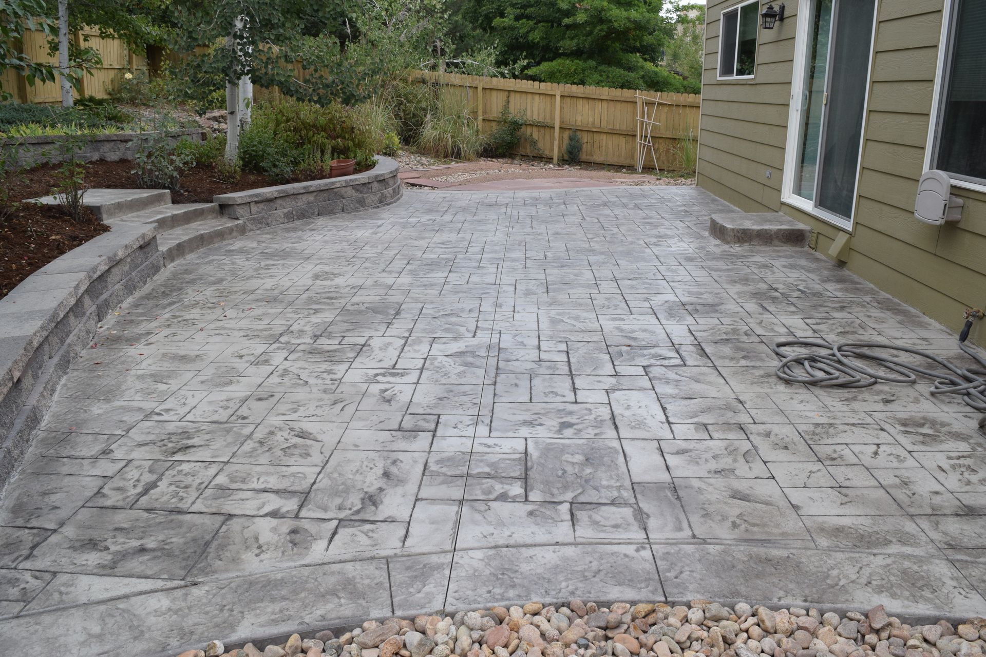 Stamped concrete patio with a gray stone pattern, next to a house and a small garden.