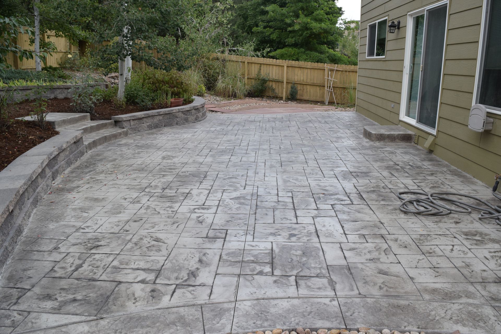 Concrete patio with textured surface next to a house and garden with a wooden fence.