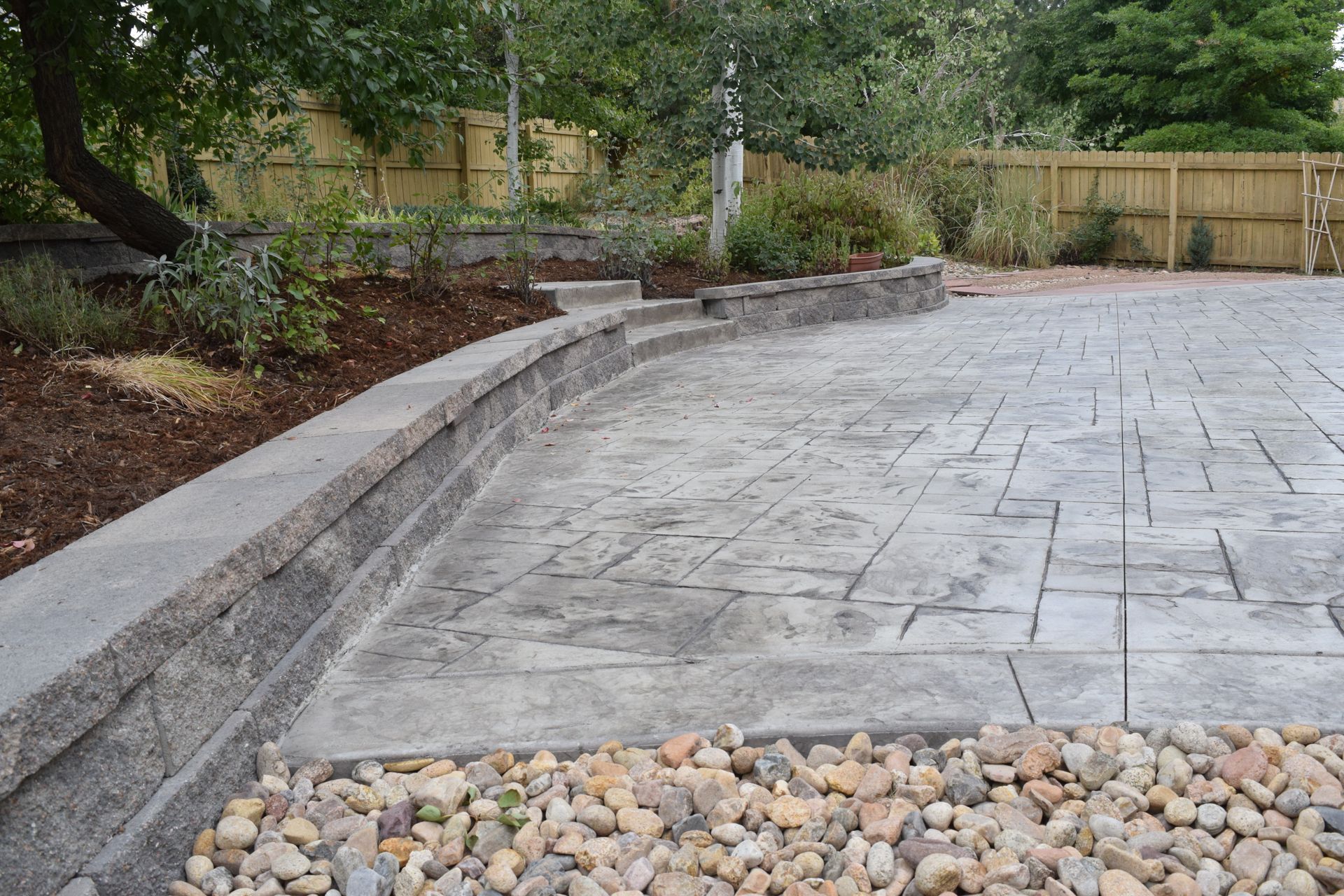 A curved stone wall borders a gray stamped concrete patio with trees and garden beds in the background.