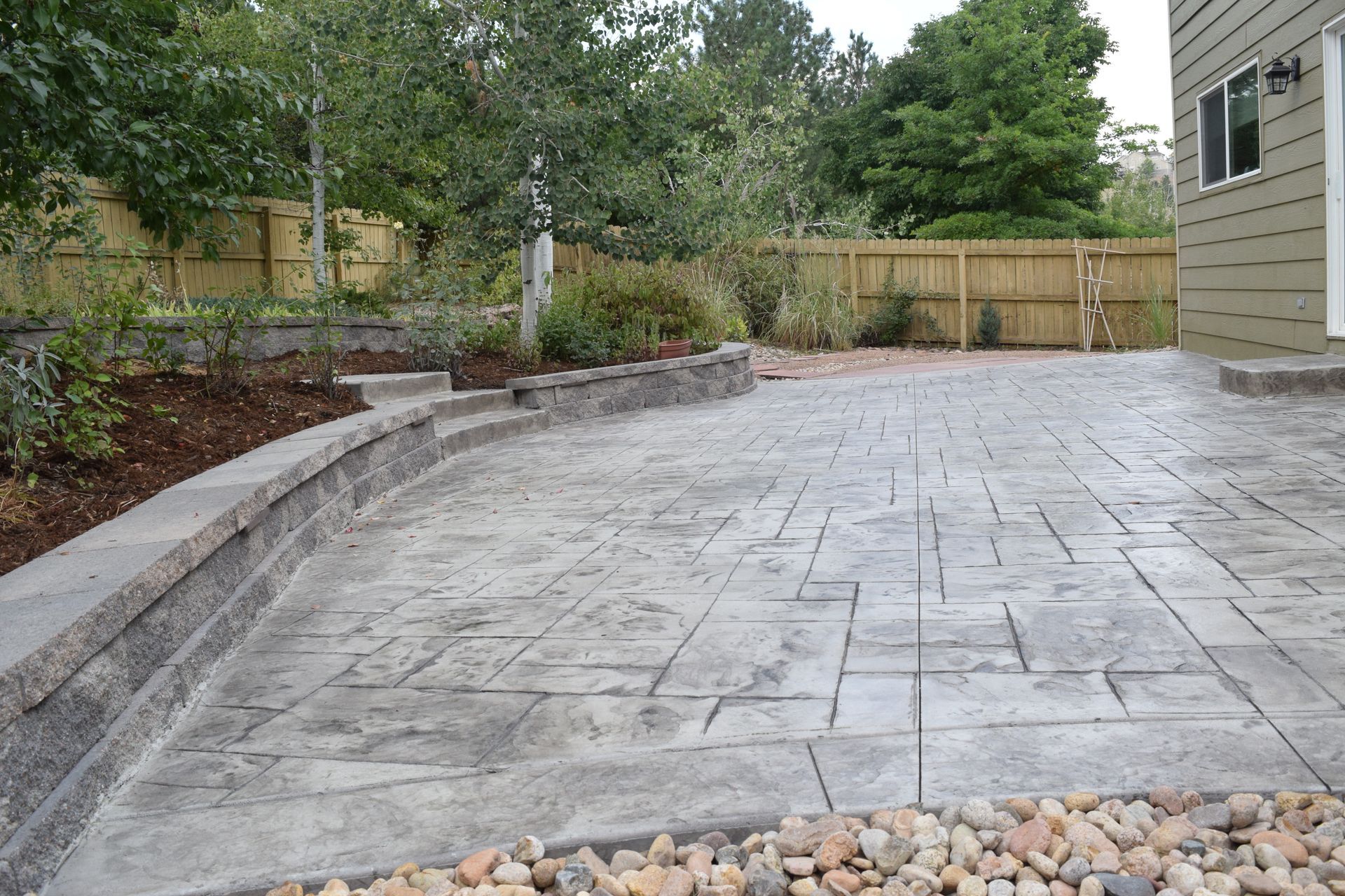 Stamped concrete patio with a retaining wall and wooden fence, with landscaping and a house exterior.