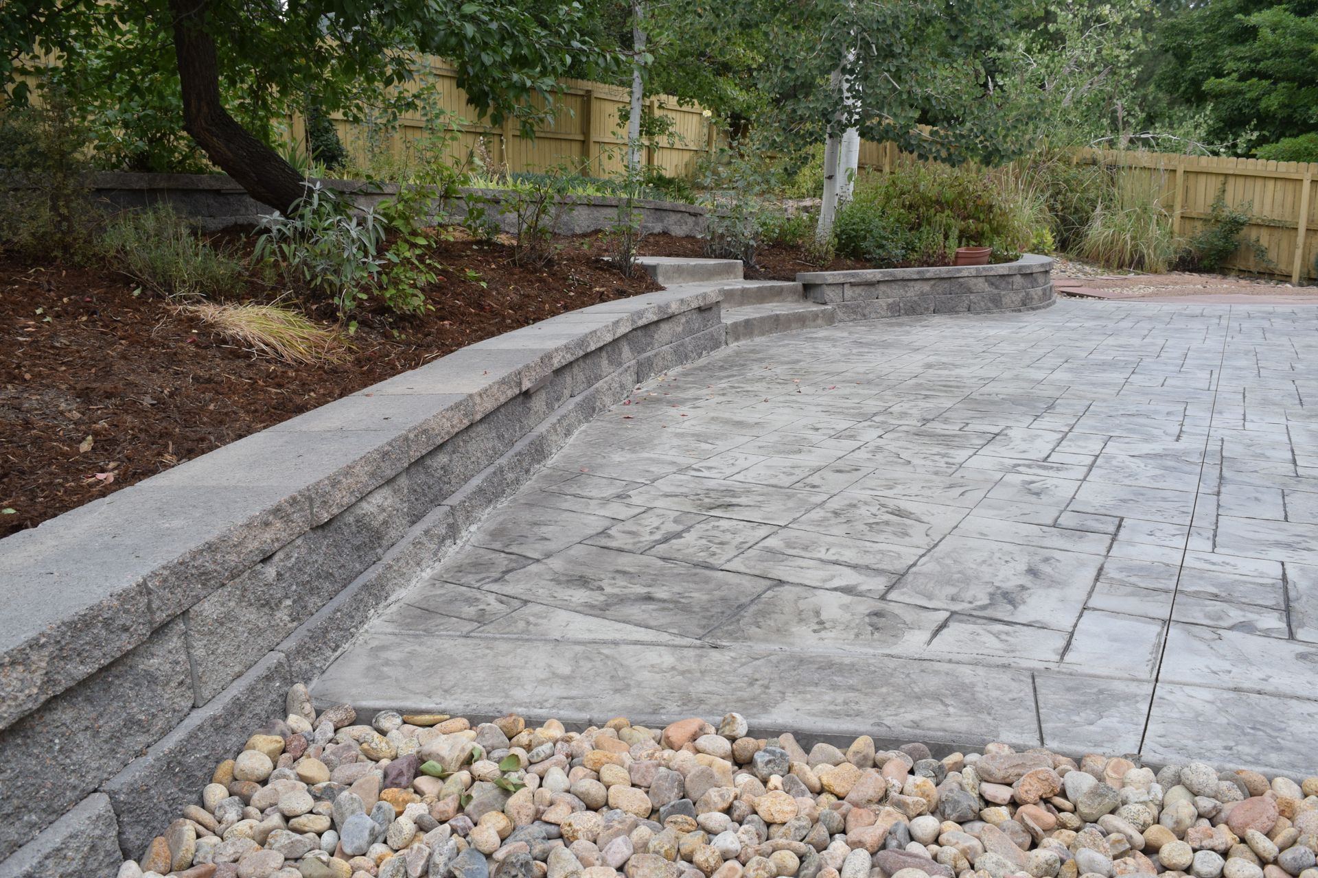 Stone patio with retaining wall, rock border, and mulch with trees and fence in background.