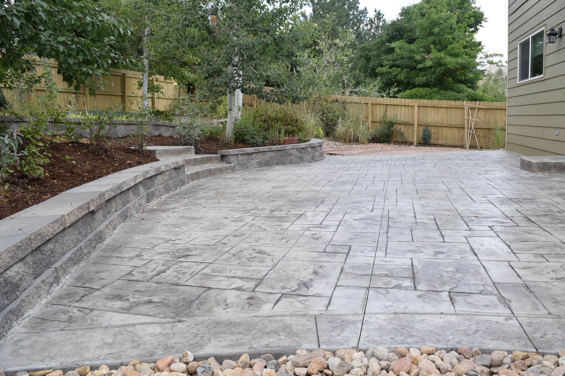Concrete patio with textured surface, bordered by a retaining wall and small gravel bed, trees in background.