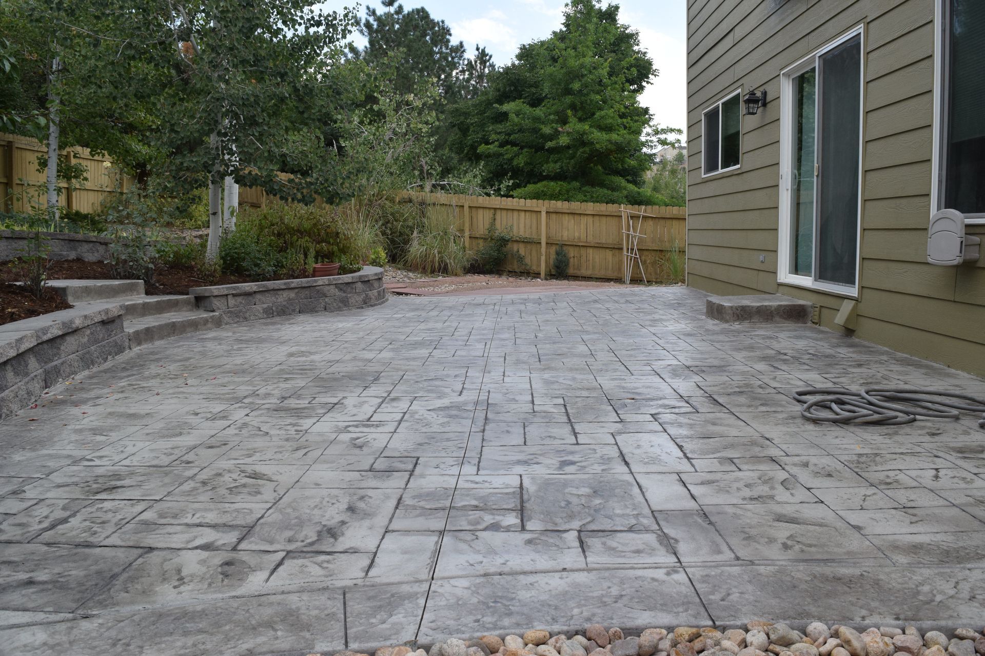Patio with gray stamped concrete, adjacent to a house. Wooden fence and trees in background.