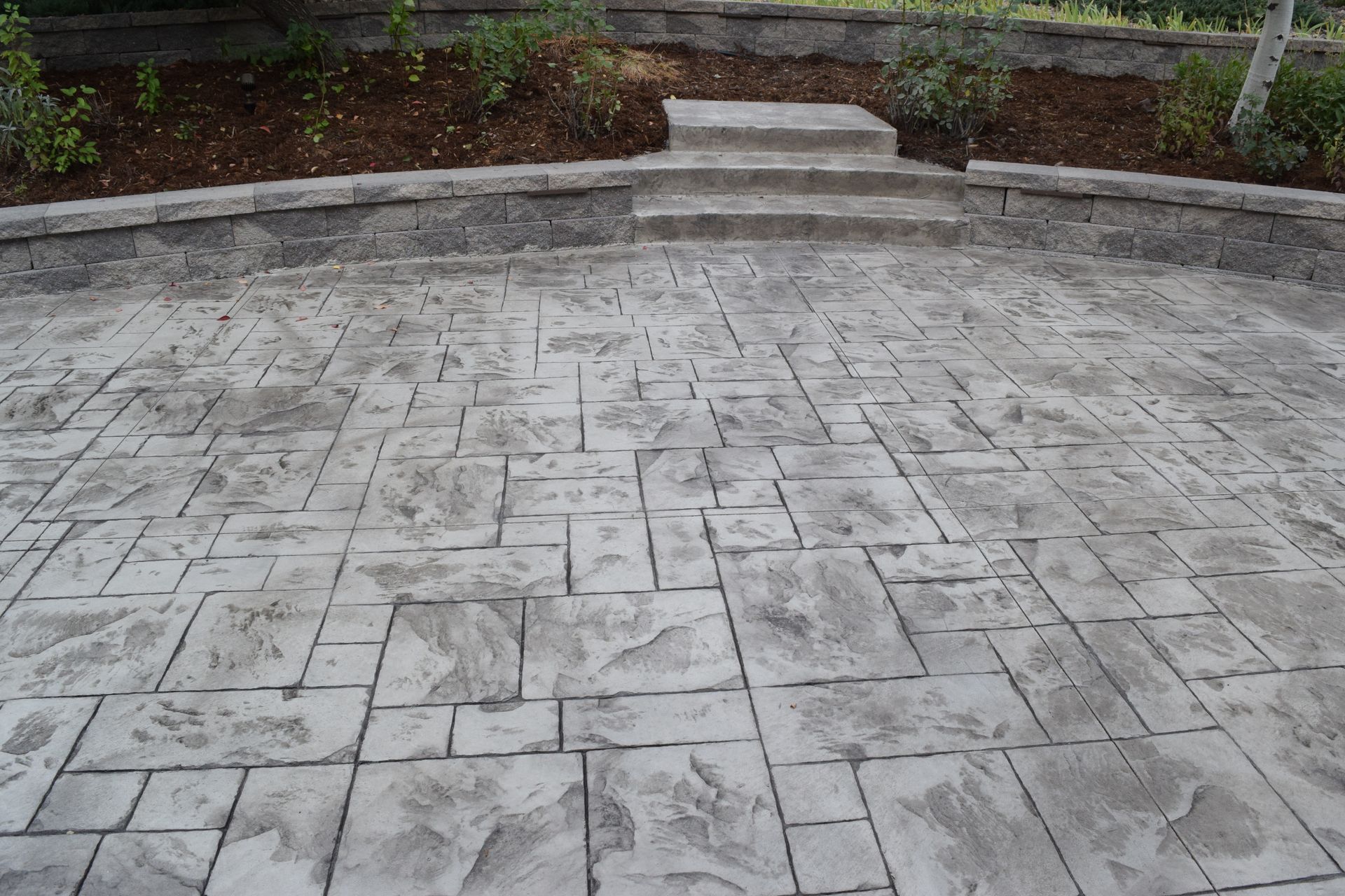 Gray stamped concrete patio with retaining wall, steps leading to a garden.