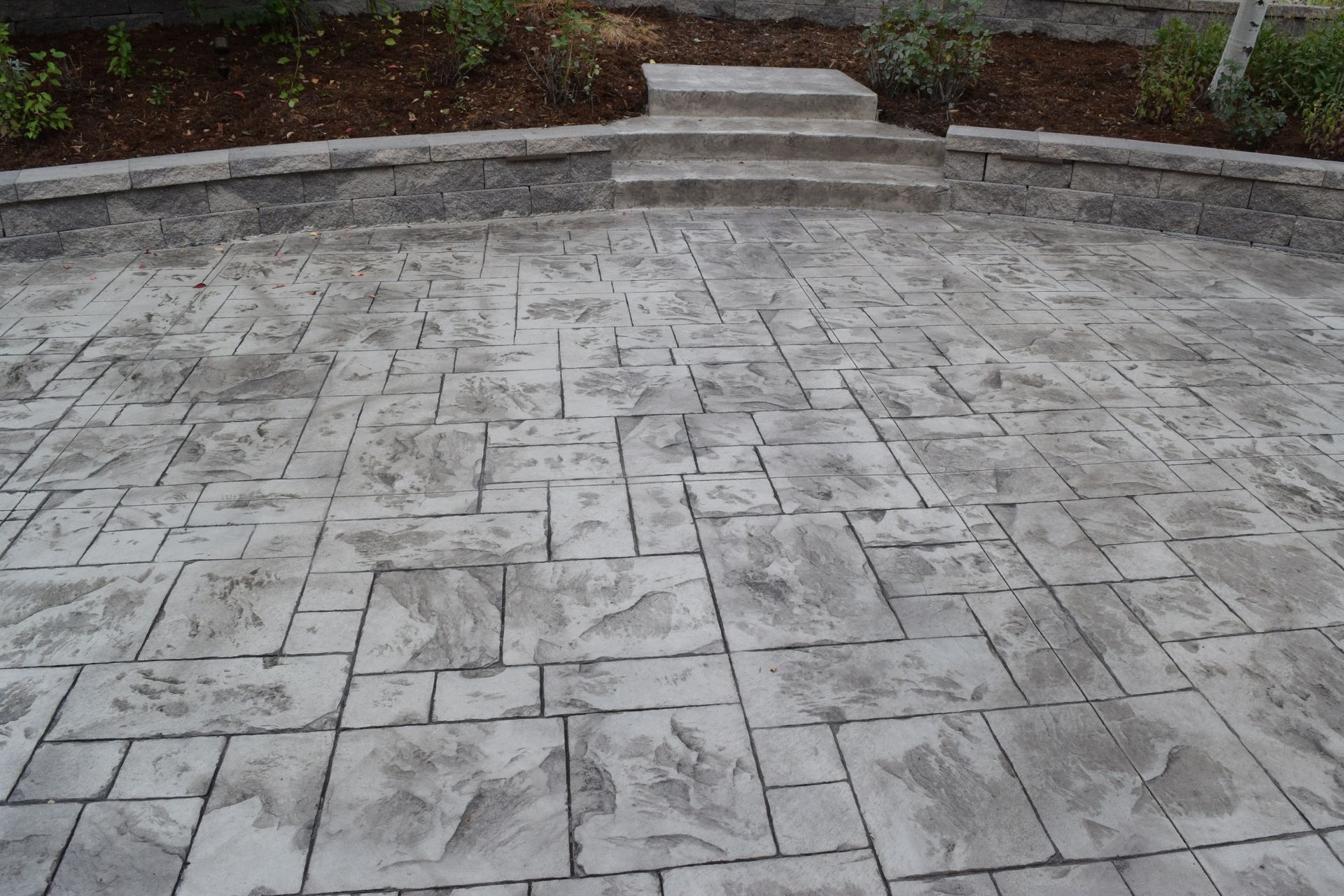 Gray stamped concrete patio with stone pattern, leading up to a raised garden bed and steps.