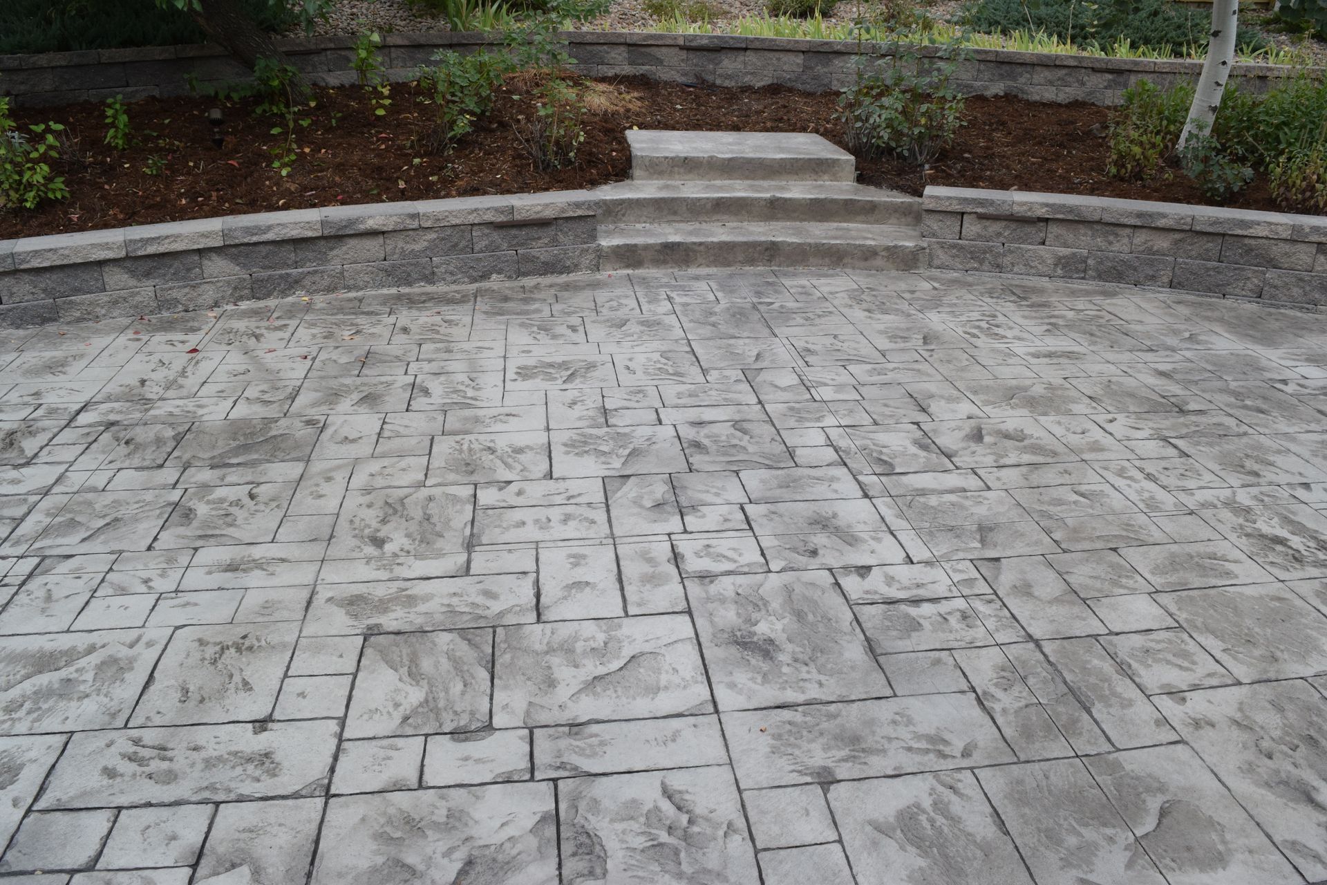 Stamped concrete patio with steps leading to a raised garden bed. Gray, stone-like texture.