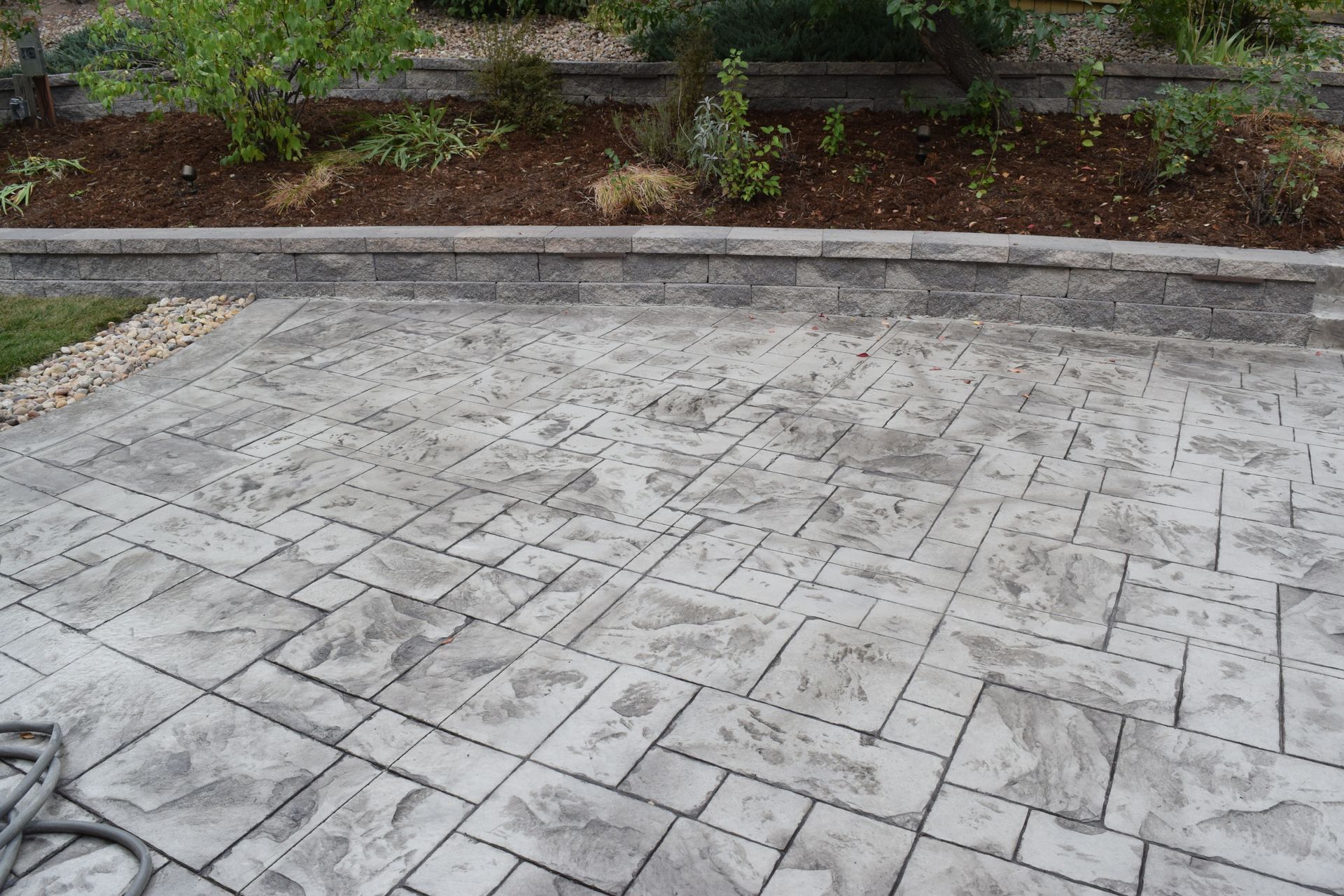 Stamped concrete patio with a gray stone pattern, bordered by a retaining wall with plants in the background.