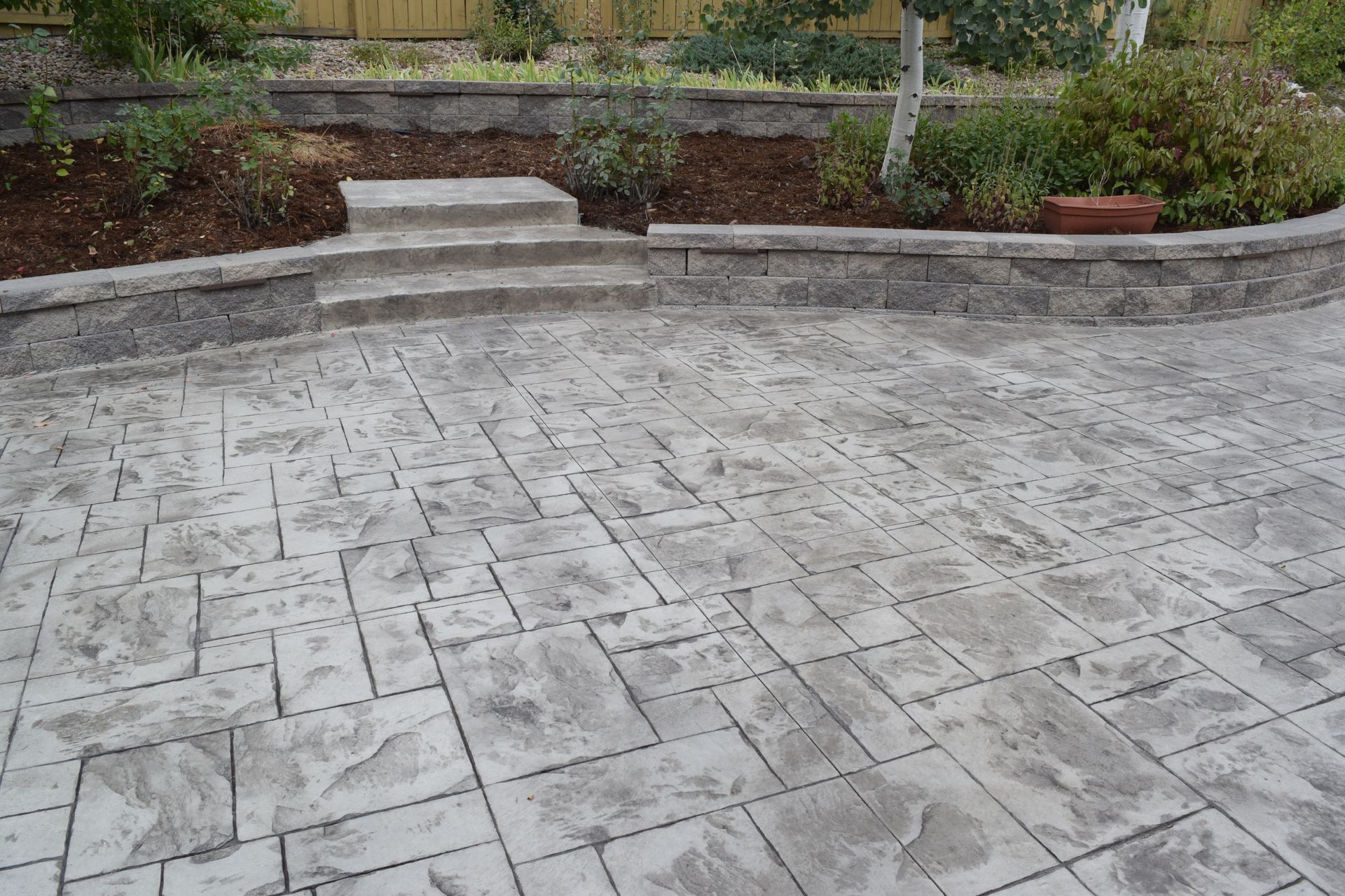 Concrete patio with a textured stone pattern, leading to steps up to a garden bed with plants and a retaining wall.