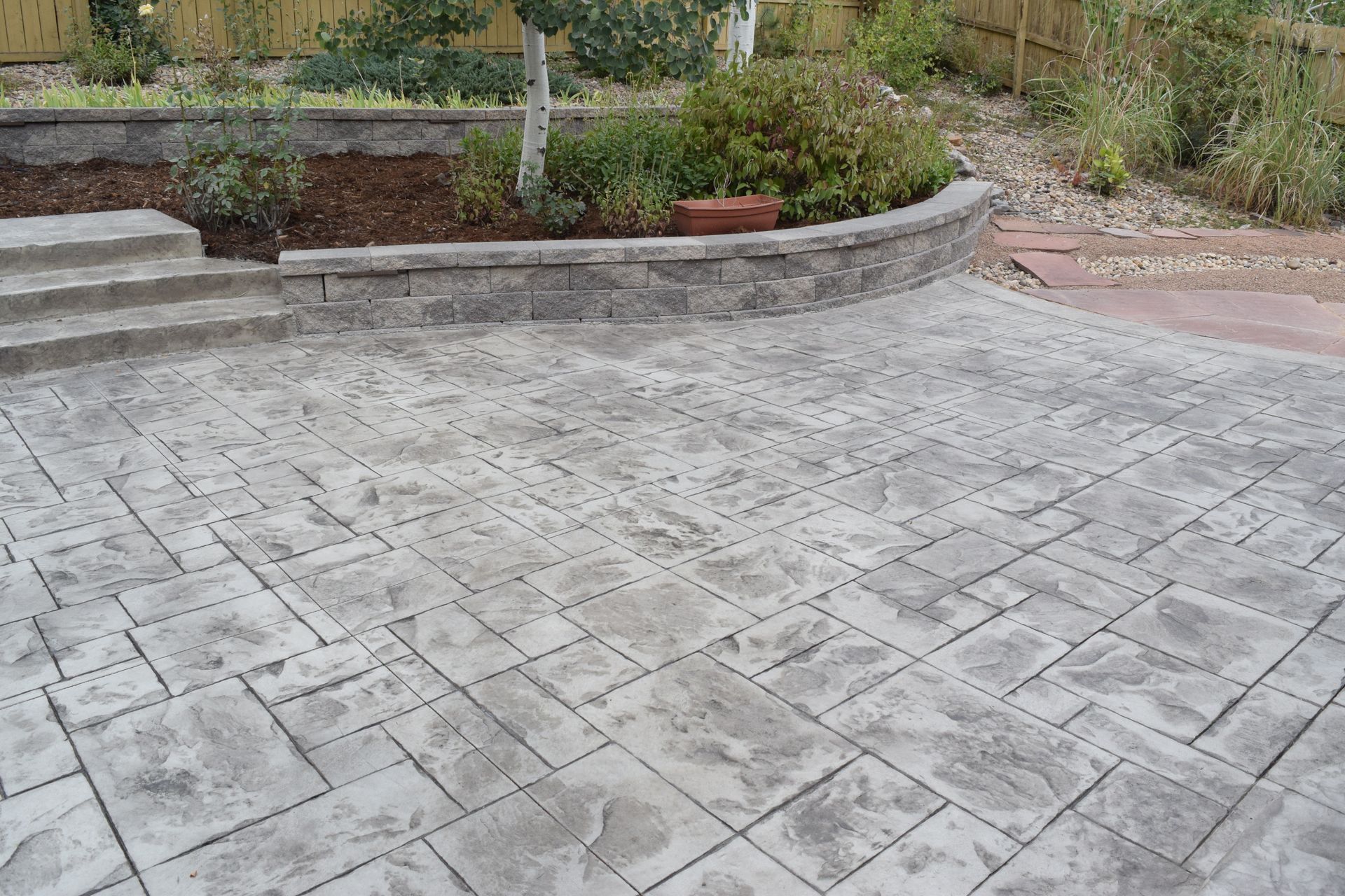 Stamped concrete patio with steps leading to a retaining wall with plants, a fence, and a brick.