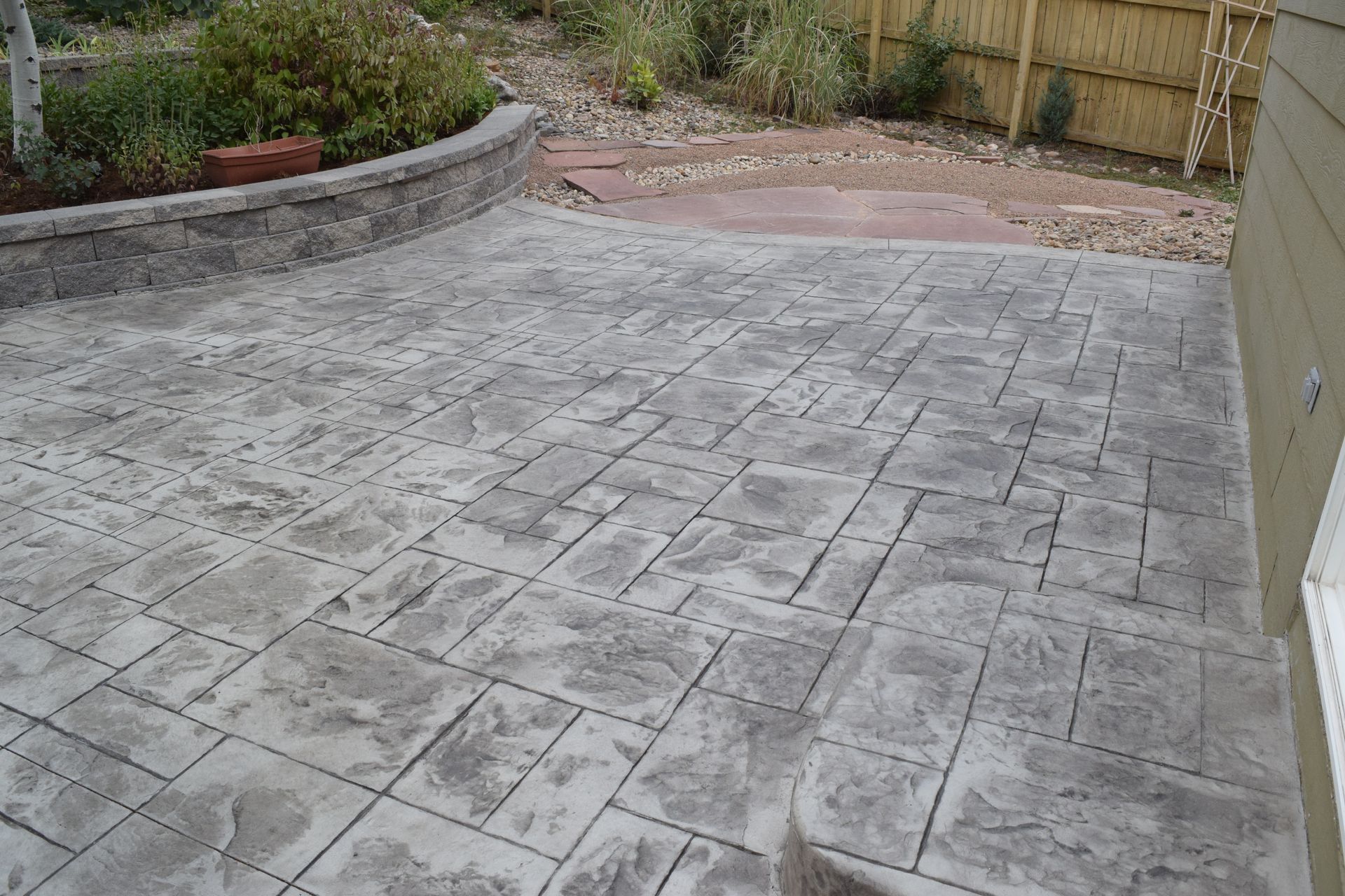 Stamped concrete patio, gray with stone pattern, next to a retaining wall and wooden fence.